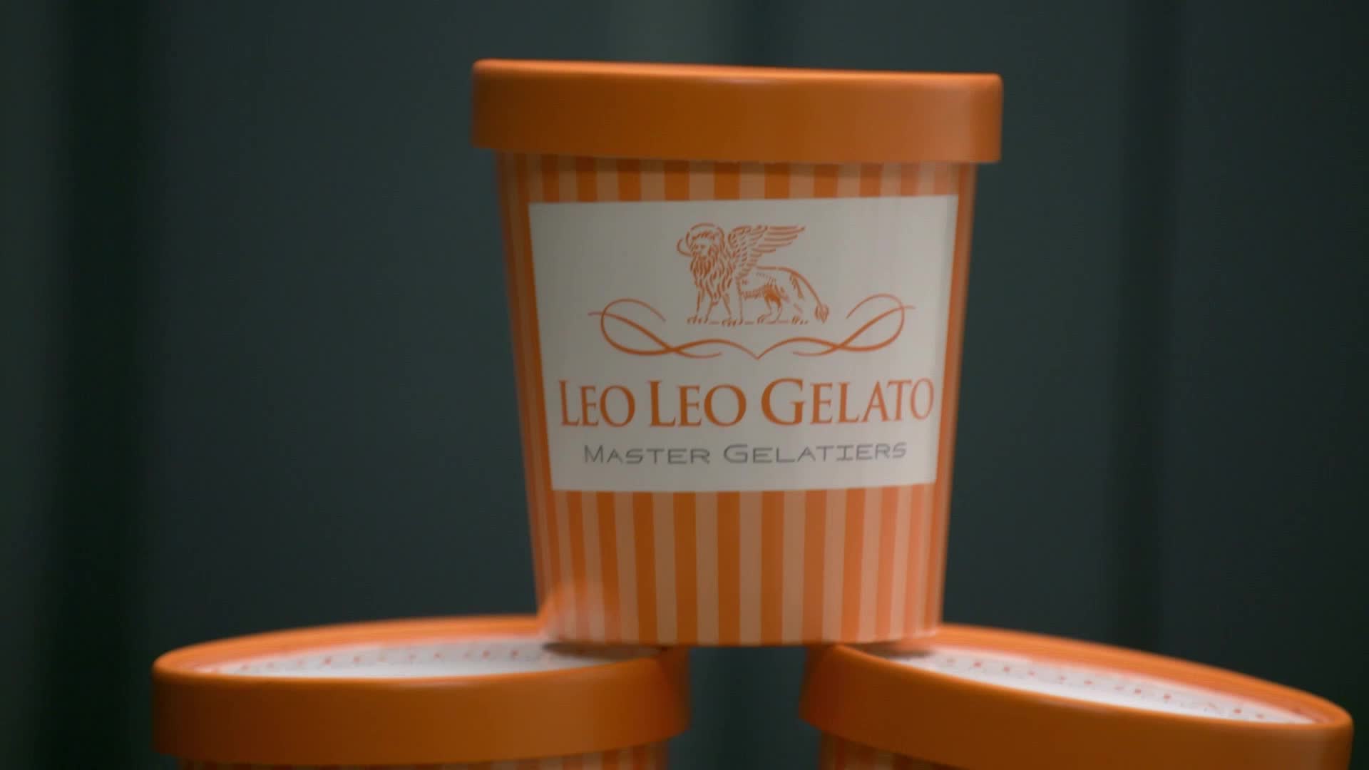 Three orange and white striped gelato containers are stacked, the top one bearing the "Leo Leo Gelato" logo. The containers' lids and the background are a darker, muted shade of teal.

