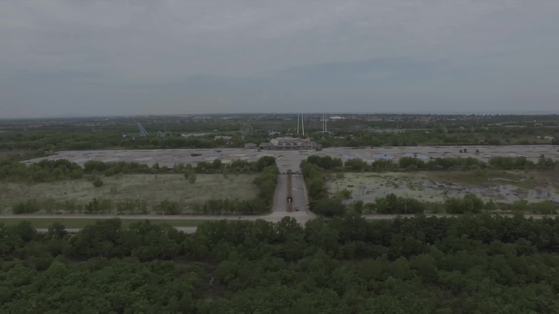 A long, empty road leads toward a large building, flanked by overgrown parking lots. The sky is overcast, and the whole scene feels still and abandoned.
