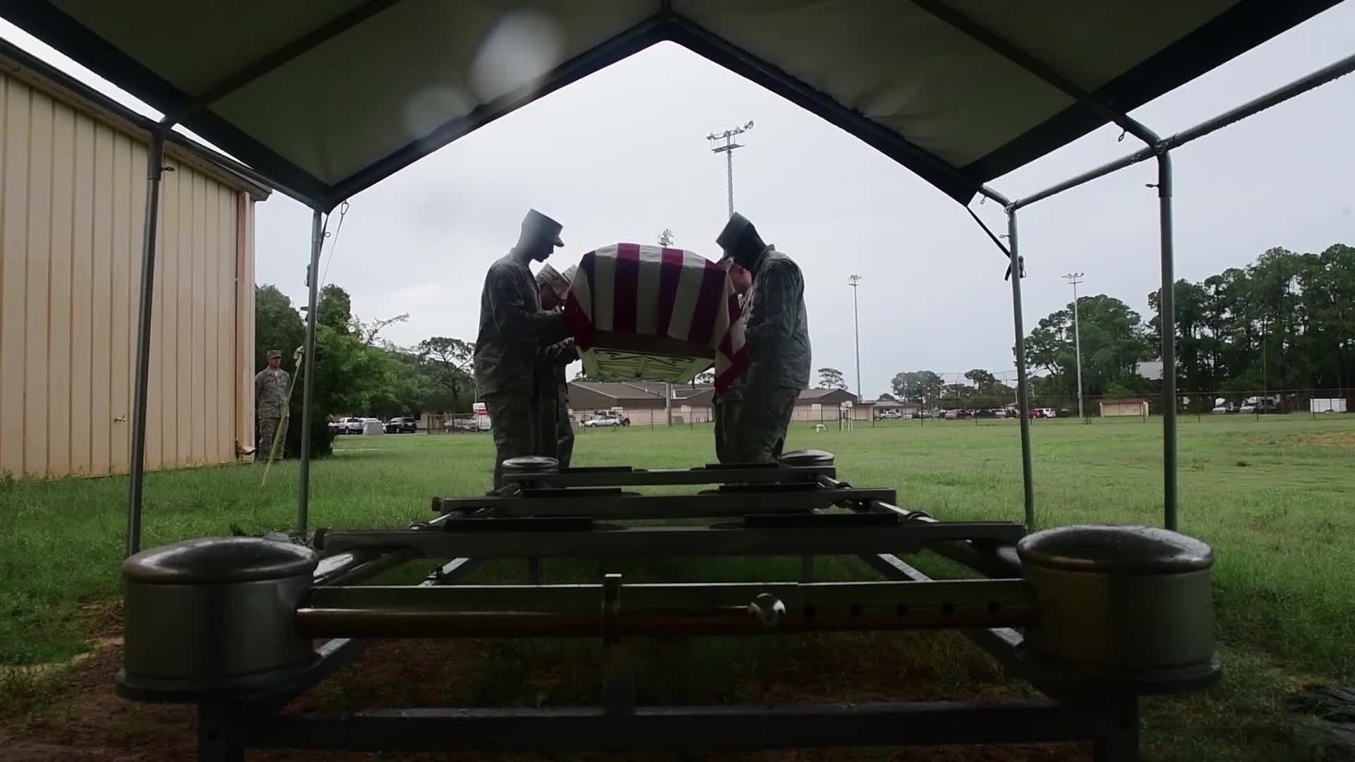 Underneath a simple canopy, three soldiers carefully carry a flag-draped casket. The somber ceremony unfolds on a damp, grassy field.
