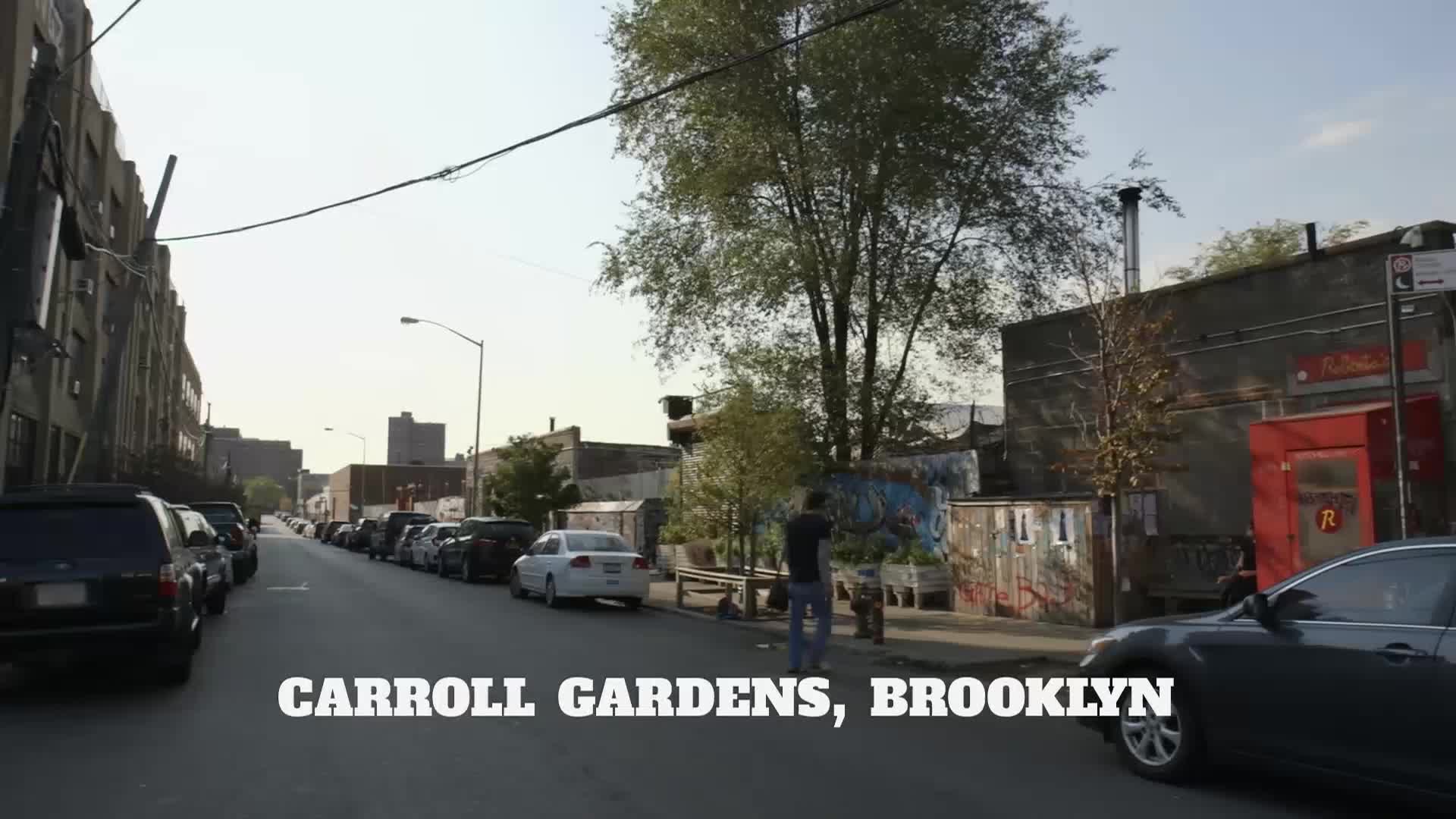 A quiet street in Carroll Gardens, Brooklyn, is lined with parked cars. A person stands near a mural, while a car drives past the camera.
