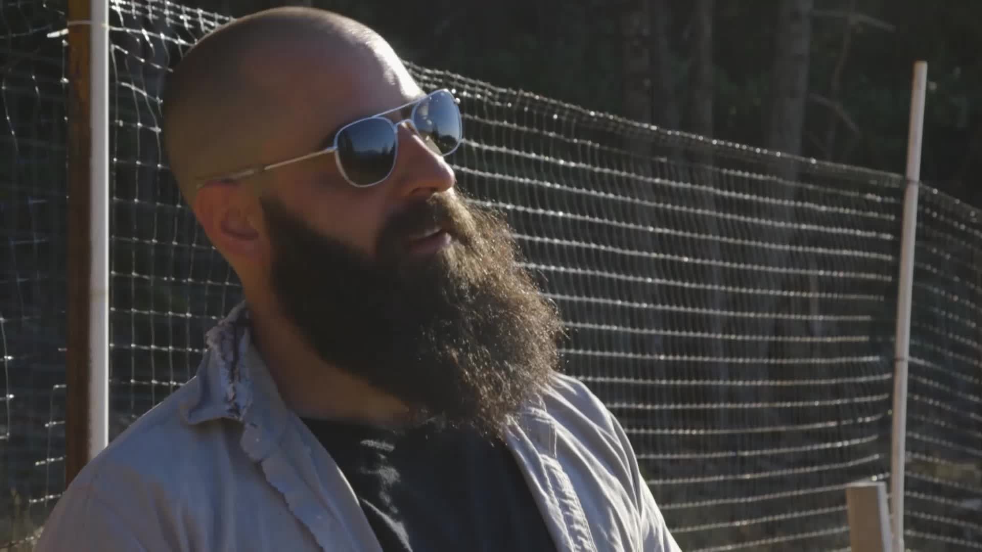 A man with a full beard and sunglasses is talking, his gaze directed slightly upward. Behind him, a wire fence and some trees are visible in the sunlight.
