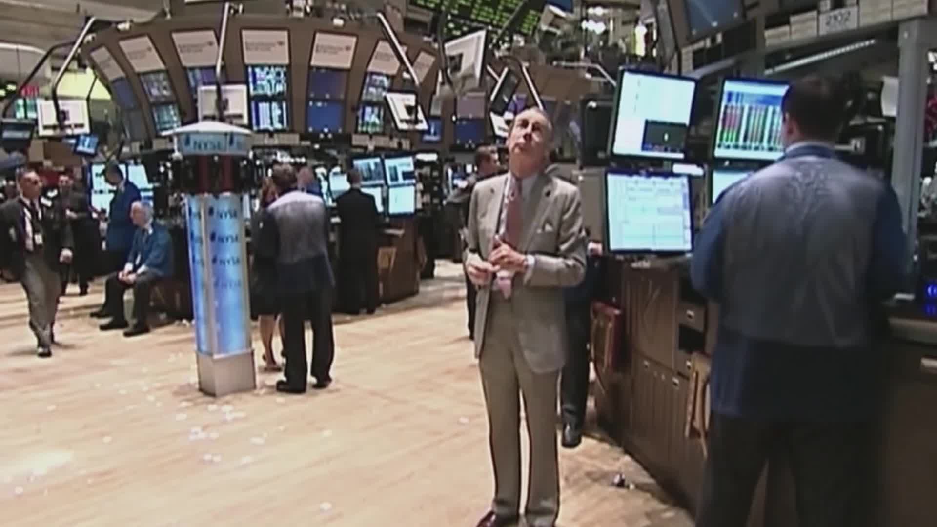 A man in a suit stands in the center, looking up as he speaks. Behind him, people mill about in a trading floor, surrounded by screens displaying data.
