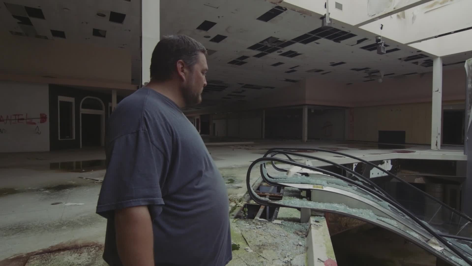 A man stands near a broken escalator, looking out over the empty space of a derelict mall. The ceiling is crumbling, and the area is dark and desolate.
A man stands near a broken escalator, looking out over the empty space of a derelict mall. The ceiling is crumbling, and the area is dark and desolate.