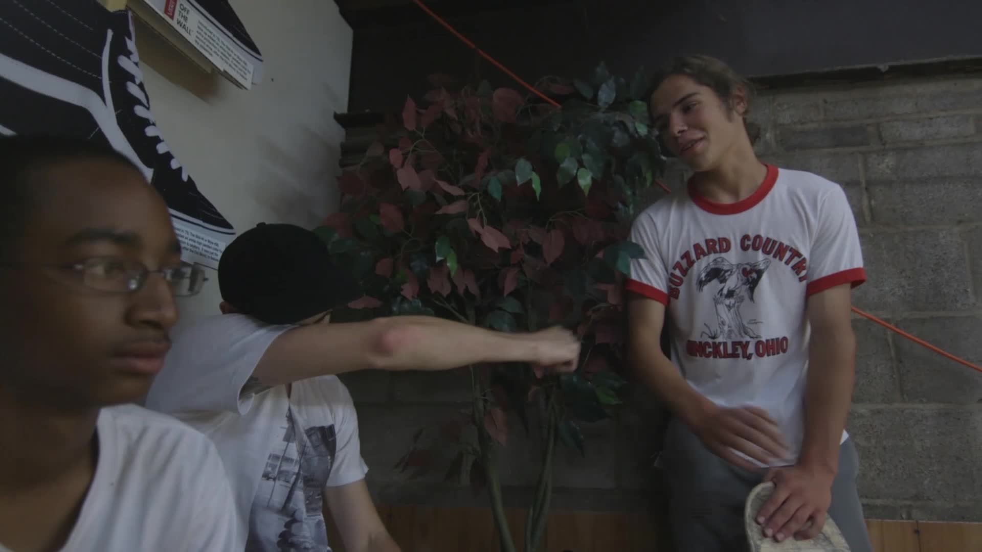 A guy in a white t-shirt throws a playful punch toward a friend. The other guy, wearing a "Buzzard County" shirt, smiles as he leans against the wall.
A guy in a white t-shirt throws a playful punch toward a friend. The other guy, wearing a "Buzzard County" shirt, smiles as he leans against the wall.