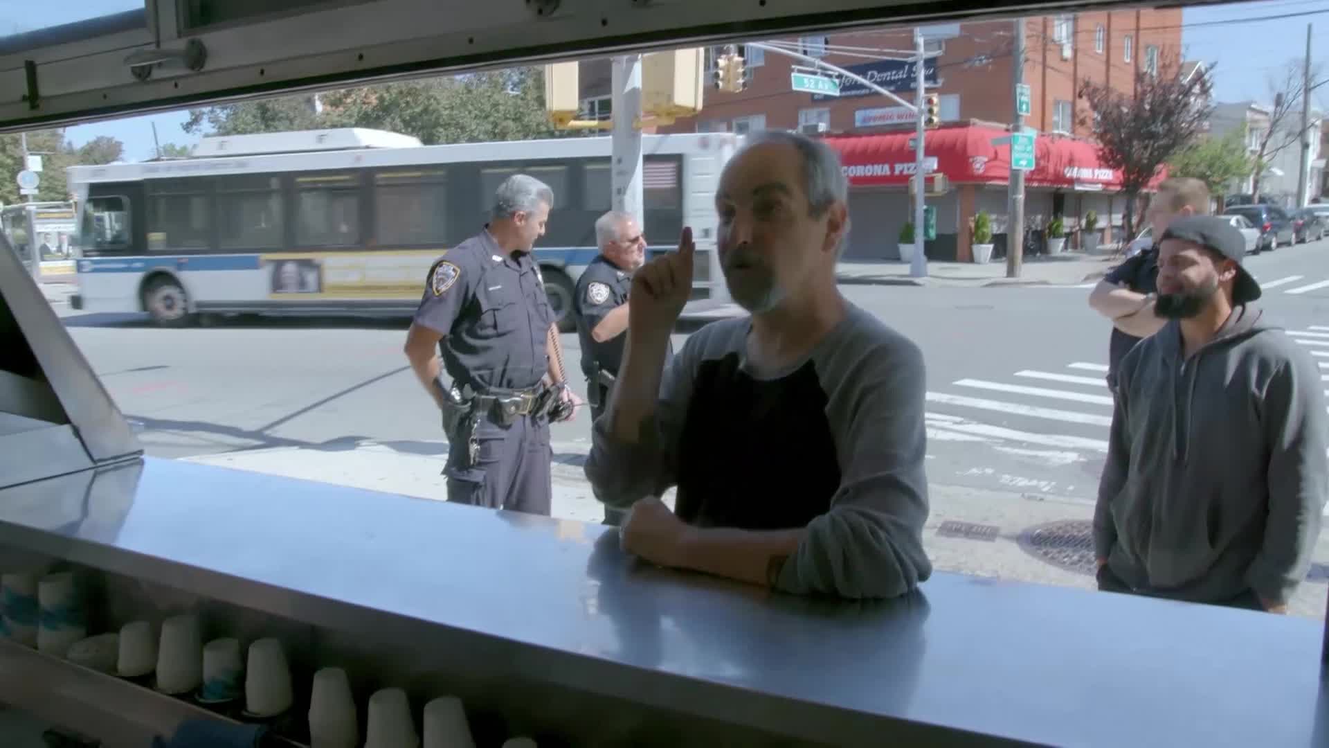 A man with graying hair gestures emphatically while talking to someone out of frame. Two uniformed police officers stand nearby, watching the interaction.
A man with graying hair gestures emphatically while talking to someone out of frame. Two uniformed police officers stand nearby, watching the interaction.