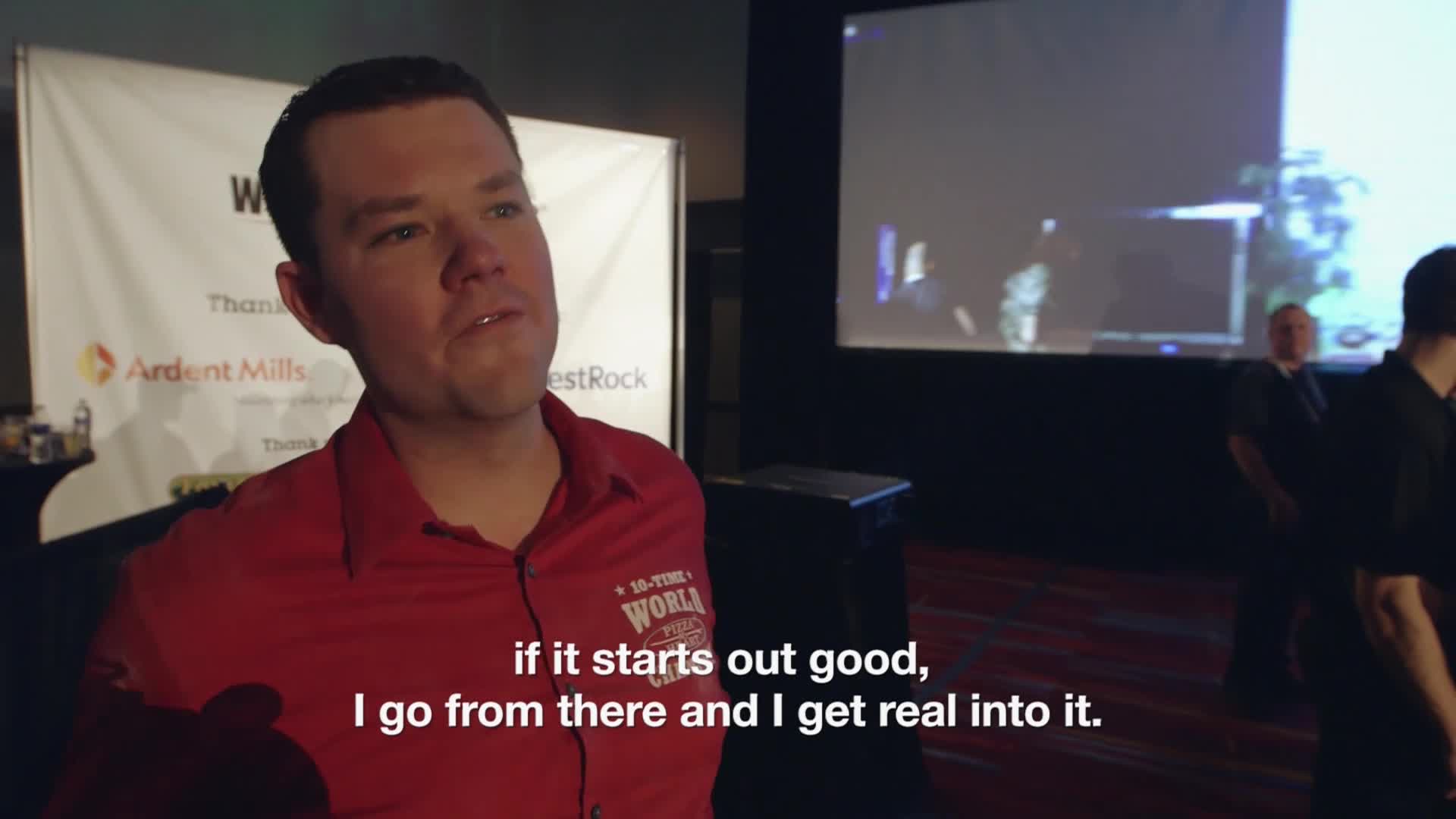 A man in a red shirt is speaking, his words subtitled on the screen. Behind him, a projection shows a blurred image as people mill around.
A man in a red shirt is speaking, his words subtitled on the screen. Behind him, a projection shows a blurred image as people mill around.
