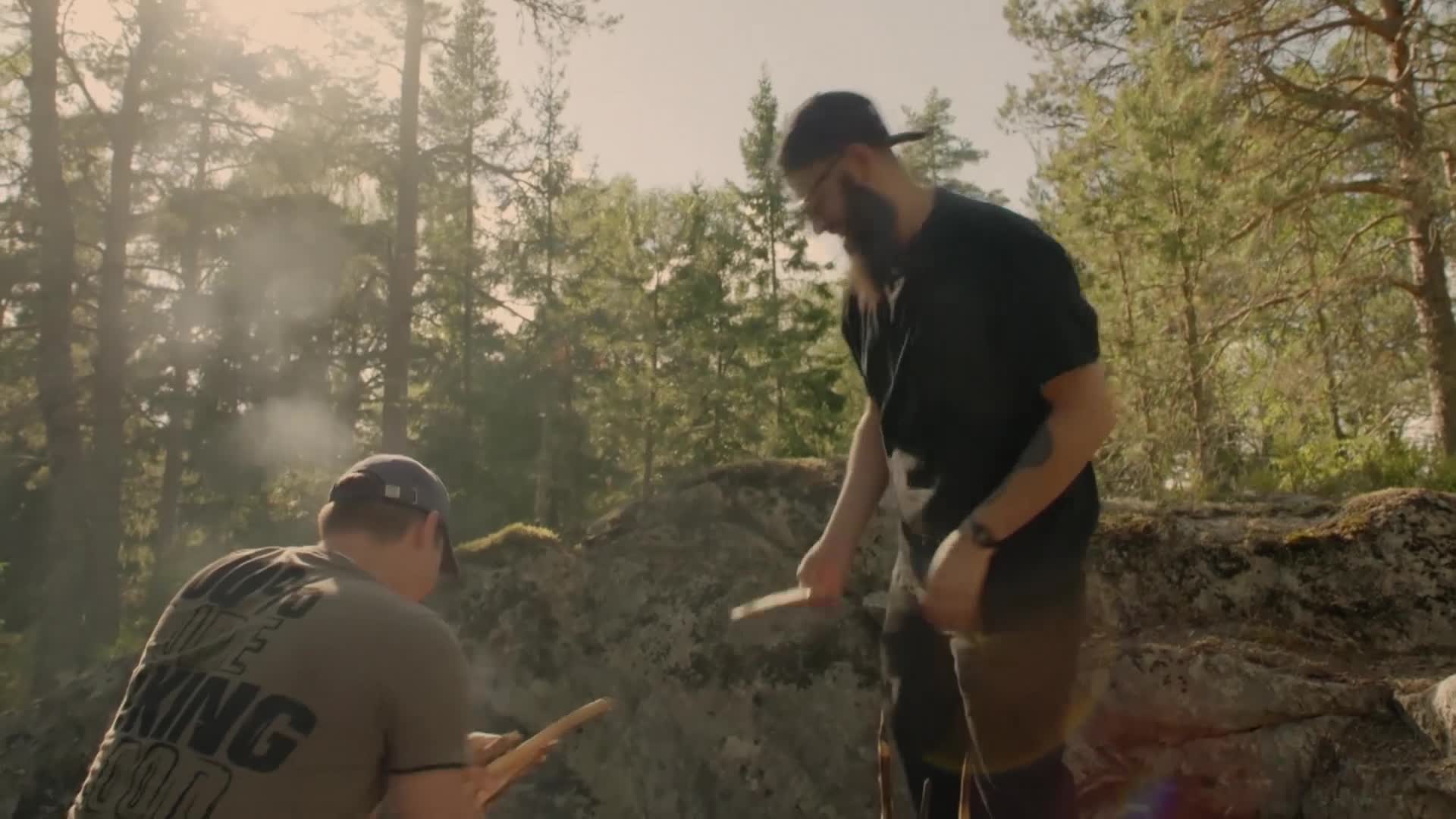 Smoke drifts up from a small fire as two men work with sticks. One, with a long beard, stands and holds a piece of wood, while the other sits, focused on his own.
Smoke drifts up from a small fire as two men work with sticks. One, with a long beard, stands and holds a piece of wood, while the other sits, focused on his own.