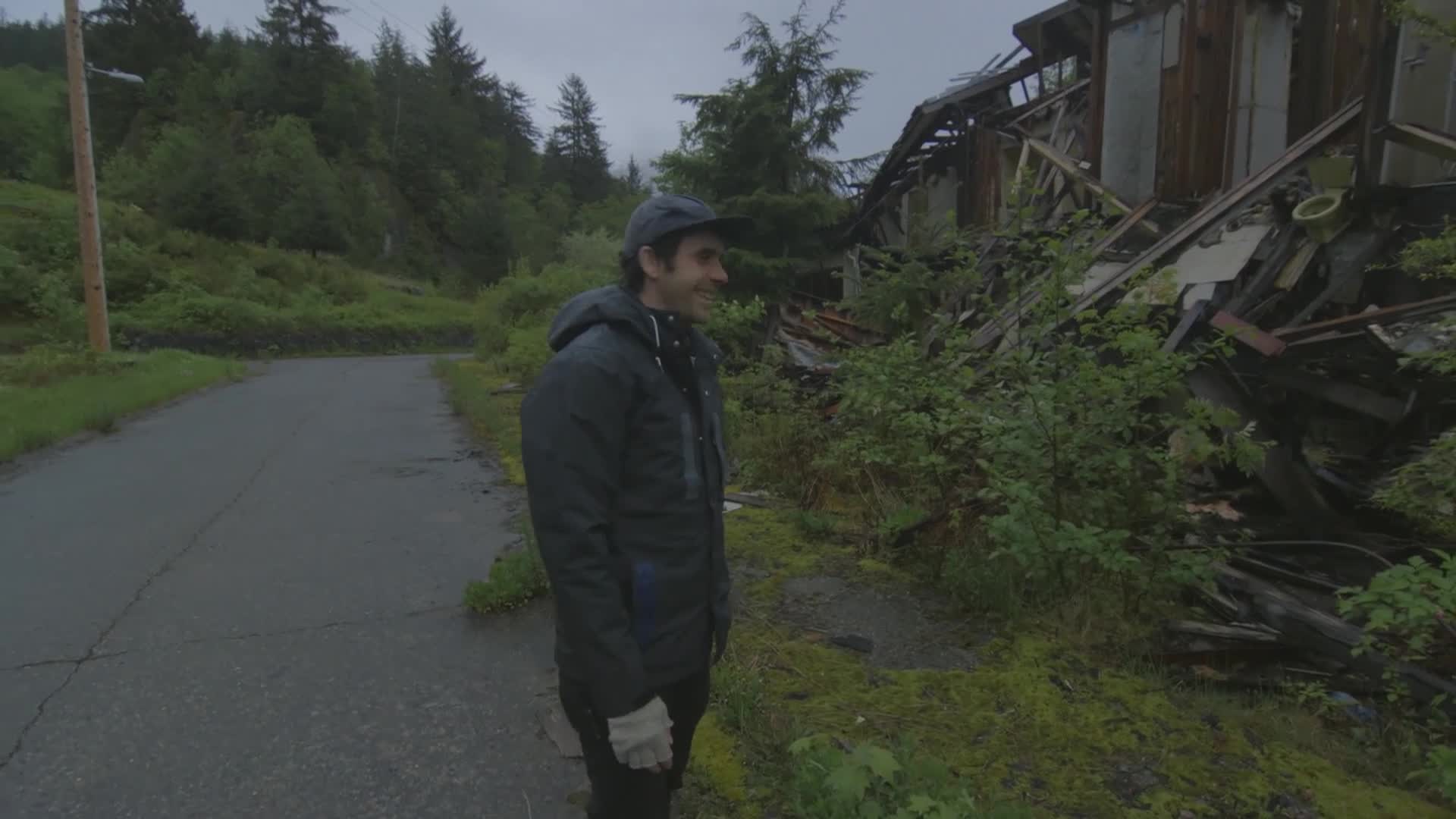 A man in a dark jacket stands on a wet road, looking toward a collapsed wooden structure overgrown with greenery. The sky is overcast, and the air feels heavy with moisture.
