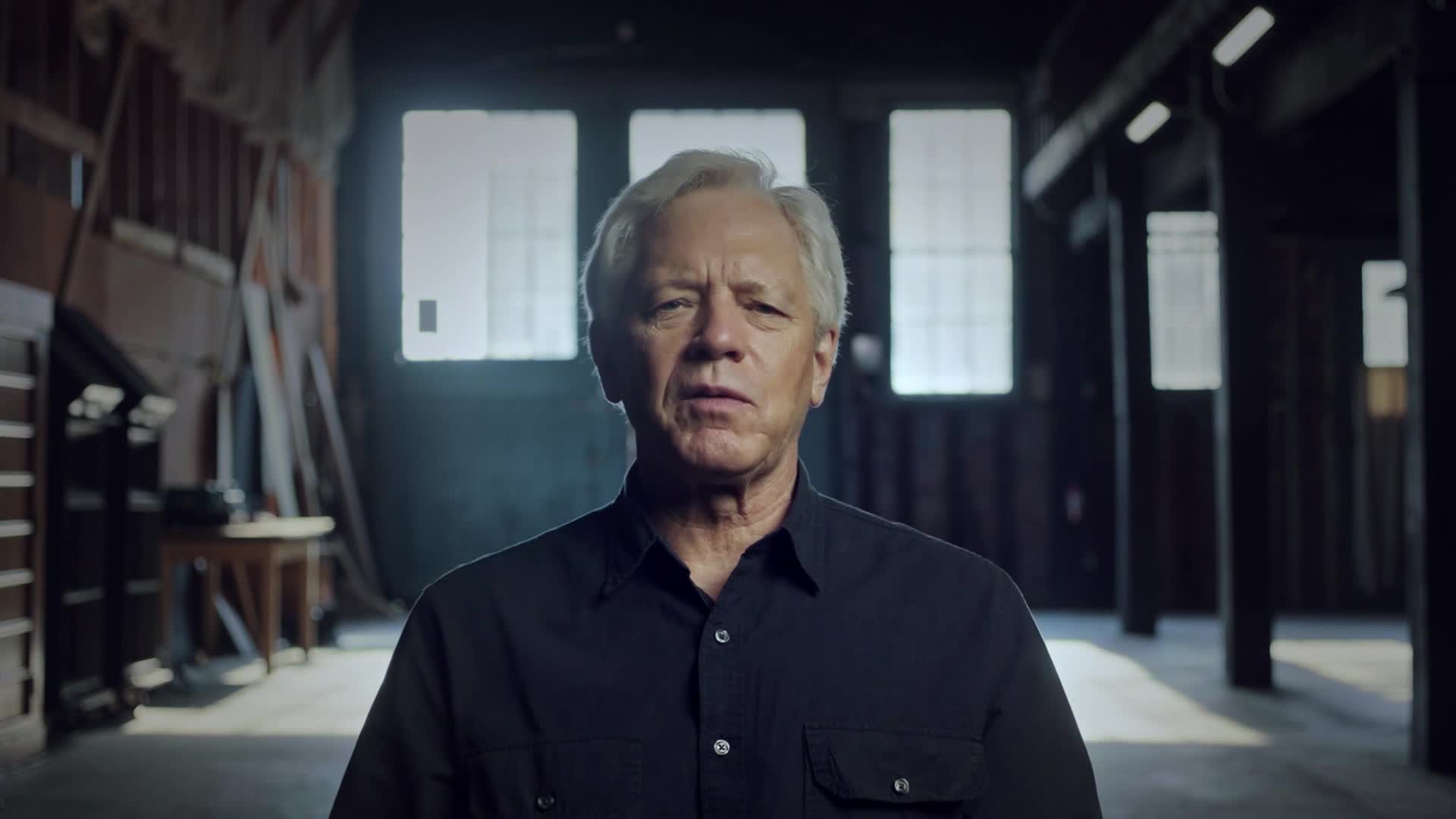 A man with silver hair and a dark shirt looks directly at the camera. He's standing in what appears to be a warehouse, with beams and windows visible in the background.
