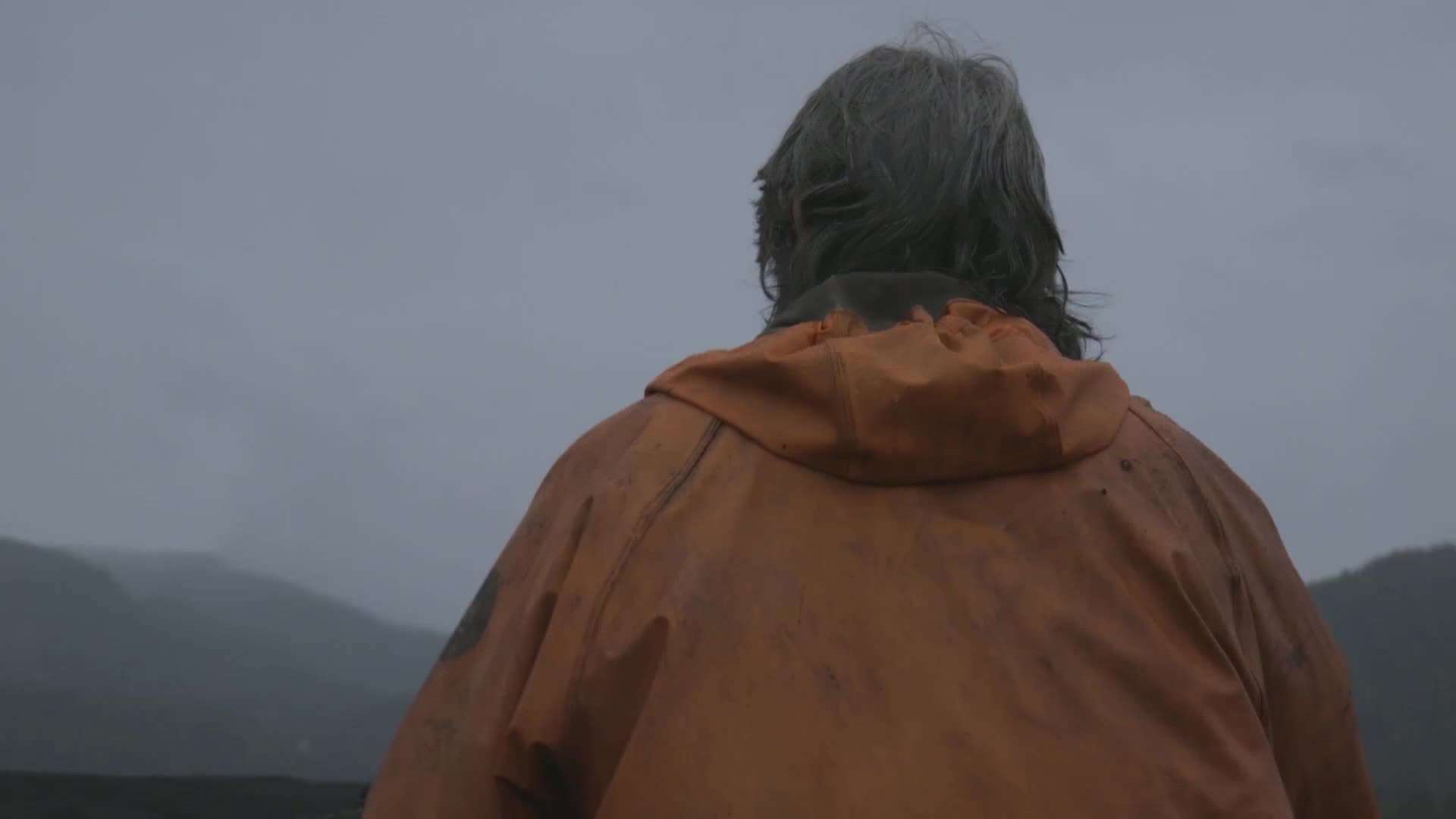 The man in the orange jacket stands facing the misty mountains. His wet, gray hair hangs down, and the back of his jacket is splattered with mud.
