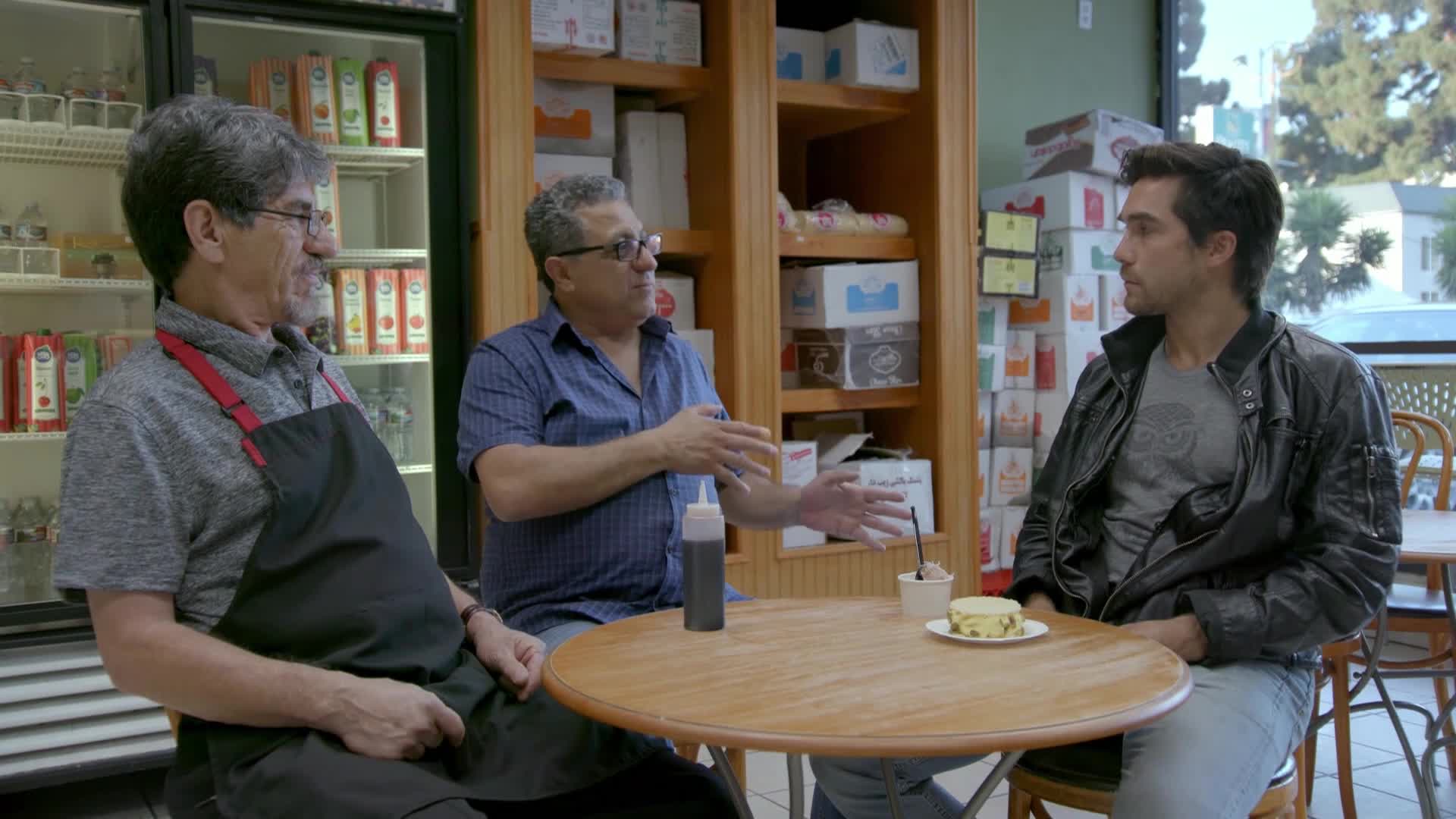 Three men sit around a small table in a shop, one gesturing animatedly with both hands. A small dessert and a bottle of sauce sit on the table between them.
