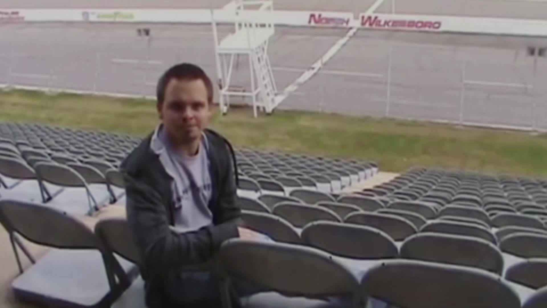 A man sits in a folding chair, looking towards the camera at the North Wilkesboro Speedway. Rows of empty seats stretch out behind him, and the track sits silent in the background.
