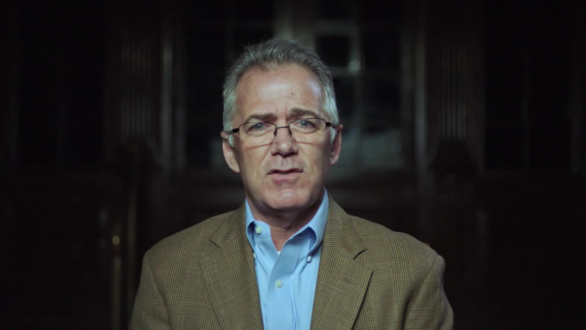 A man in a blazer and glasses is speaking directly to the camera, his expression serious. The background is dim, with dark, wooden architectural details barely visible.
