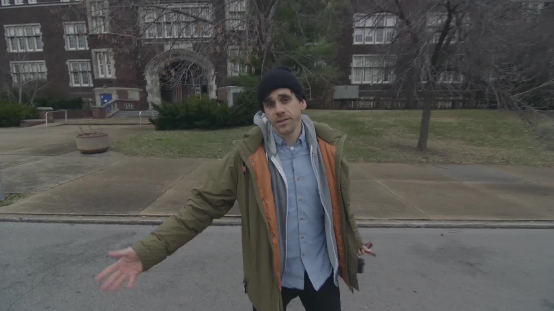 A man in a beanie and jacket stands on a street in front of a brick building. He gestures with his hands, as if explaining something for a VICE TV segment.
