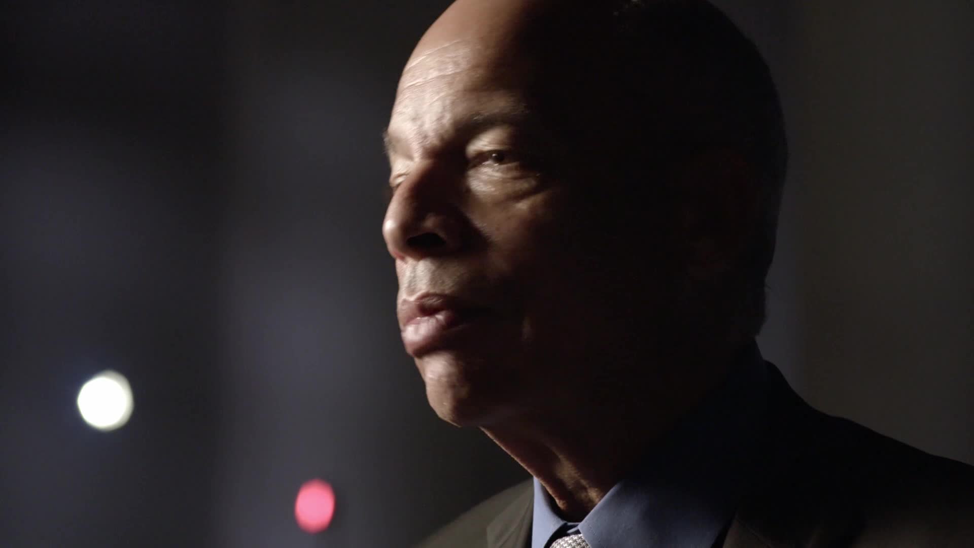 A man with a shaved head is speaking, his lips slightly parted. The background is dark, with a few blurred lights visible.
