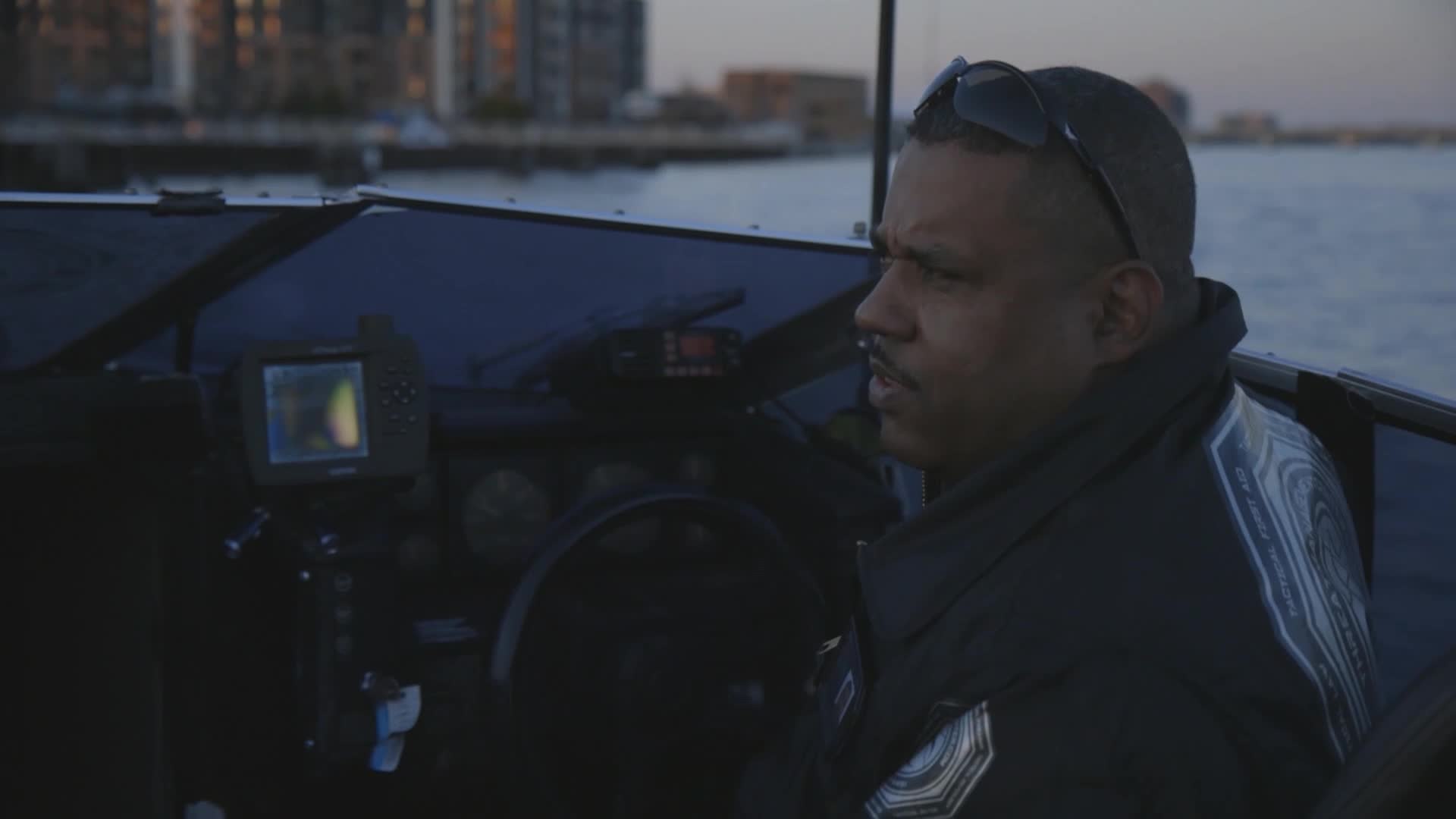 A man in a dark uniform sits at the helm of a boat, looking off to the side. The screen of a GPS device is lit up on the dashboard.
