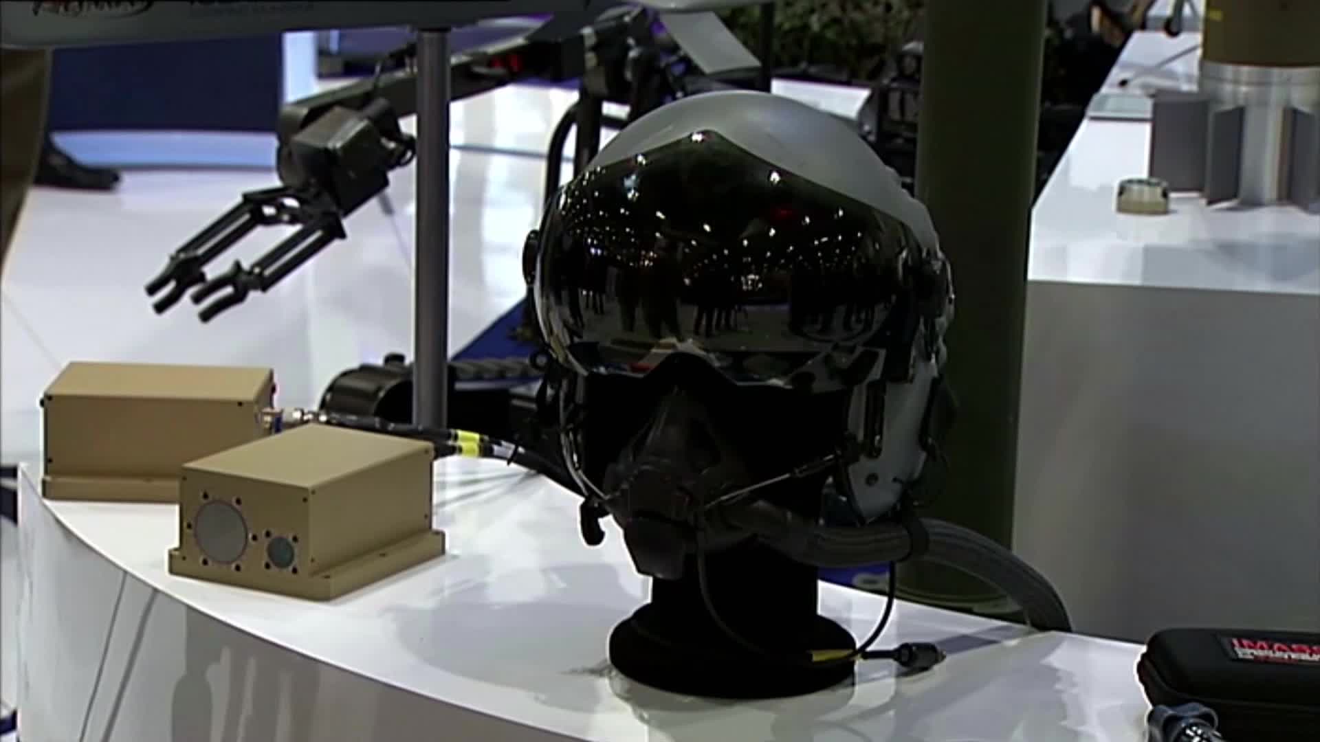 A pilot's helmet with a dark visor sits on a black stand, reflecting the surrounding environment. Two beige boxes and a robotic arm are displayed nearby, likely part of an exhibit.
