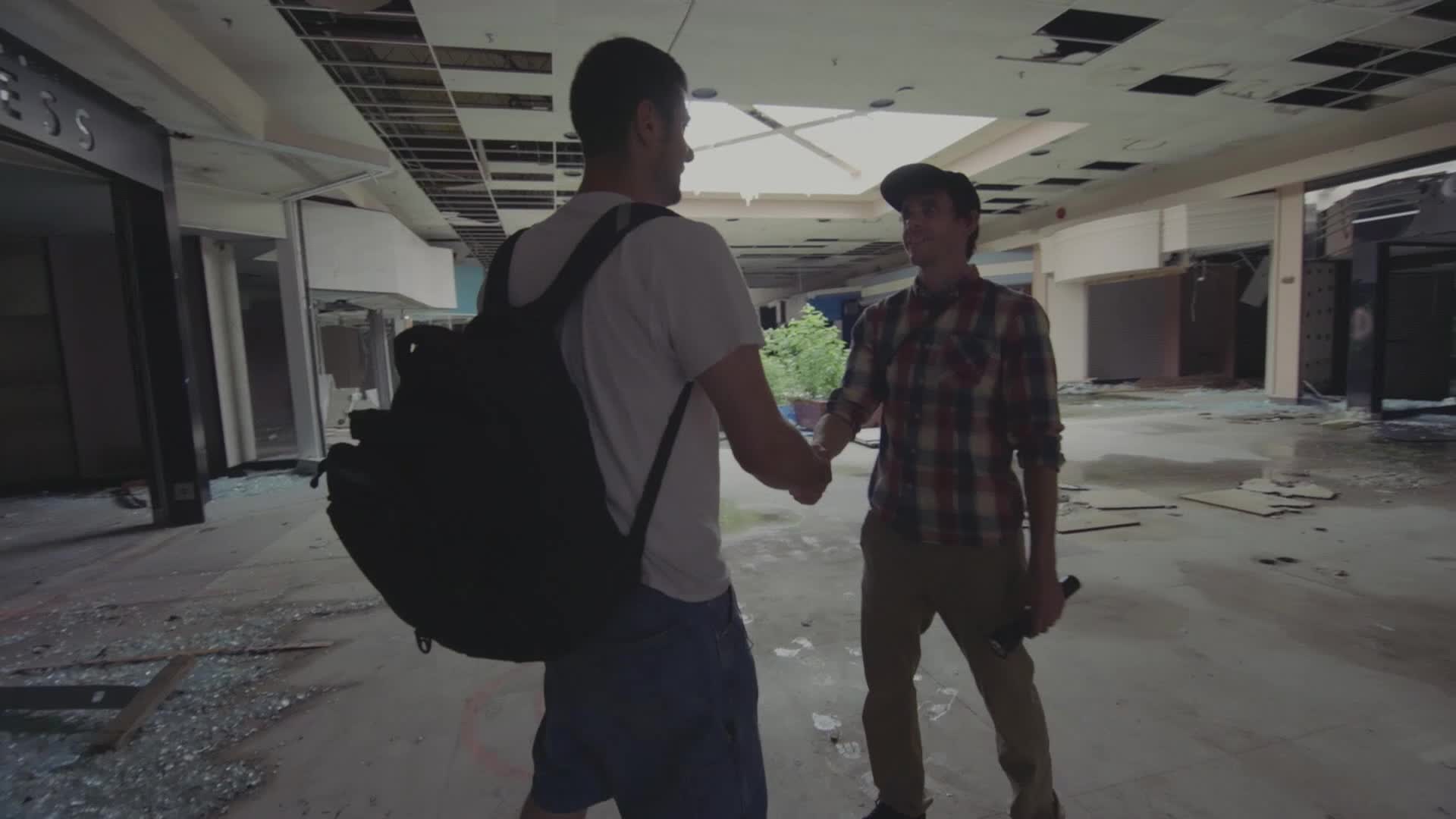 Two guys shake hands in the middle of a gutted-out mall, the ceiling partially collapsed. One of them is wearing a backpack, and the other holds a dark object.
