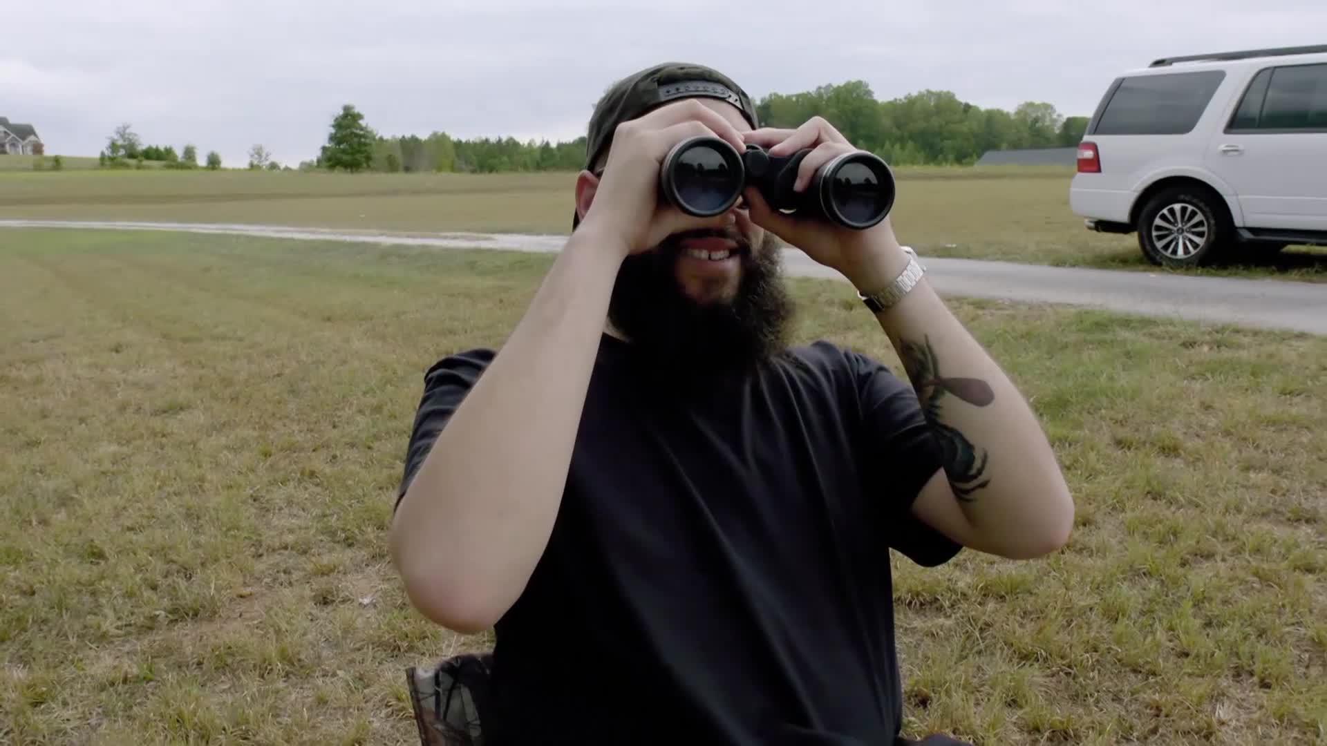 A man with a beard and a backwards cap is sitting in a chair, peering through binoculars. Behind him, a white SUV sits on a gravel road, and the landscape stretches out toward a distant tree line.

