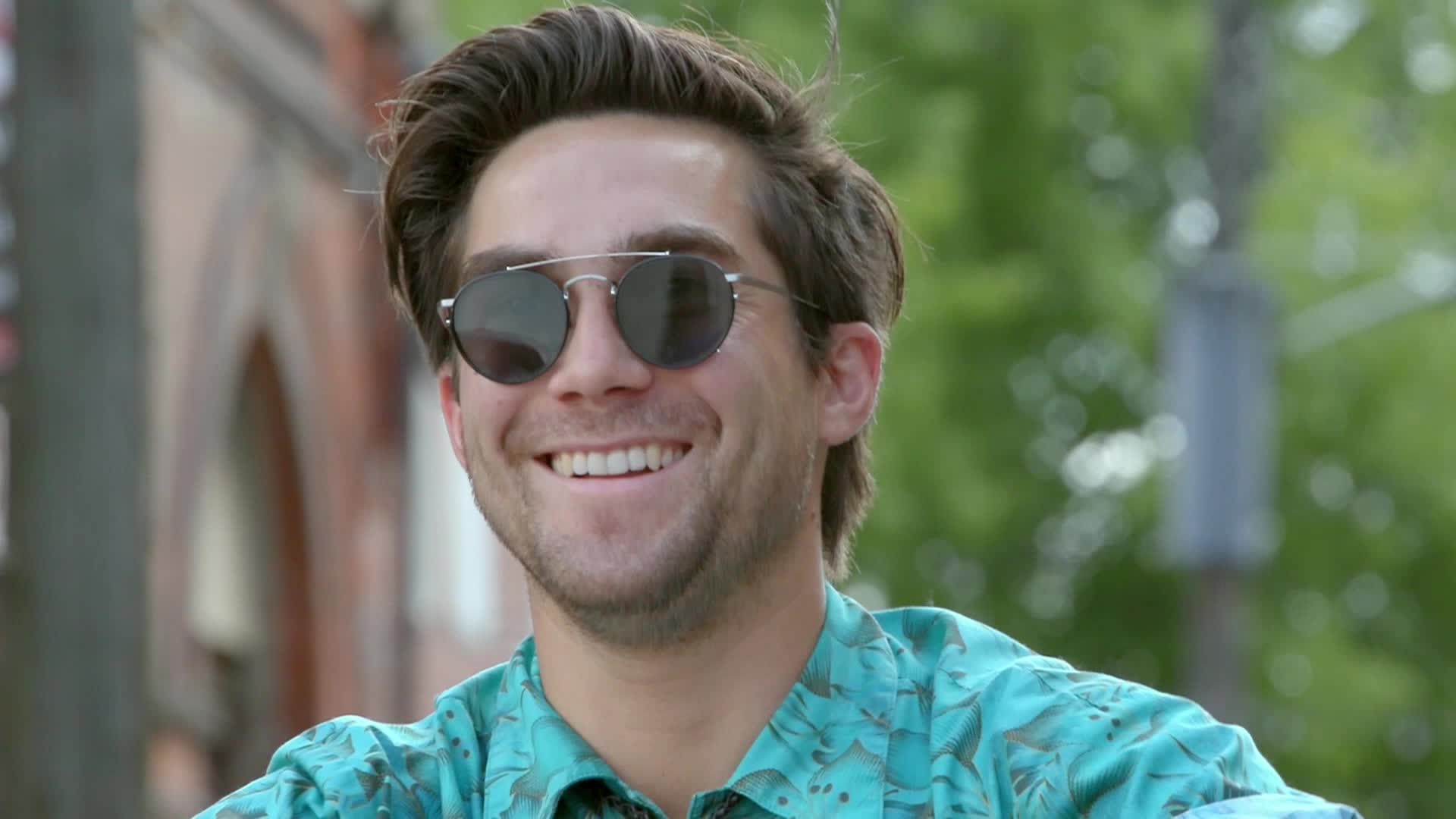 The man in sunglasses is grinning, his teeth on full display. He's wearing a patterned shirt, and the background is blurred with greenery.
