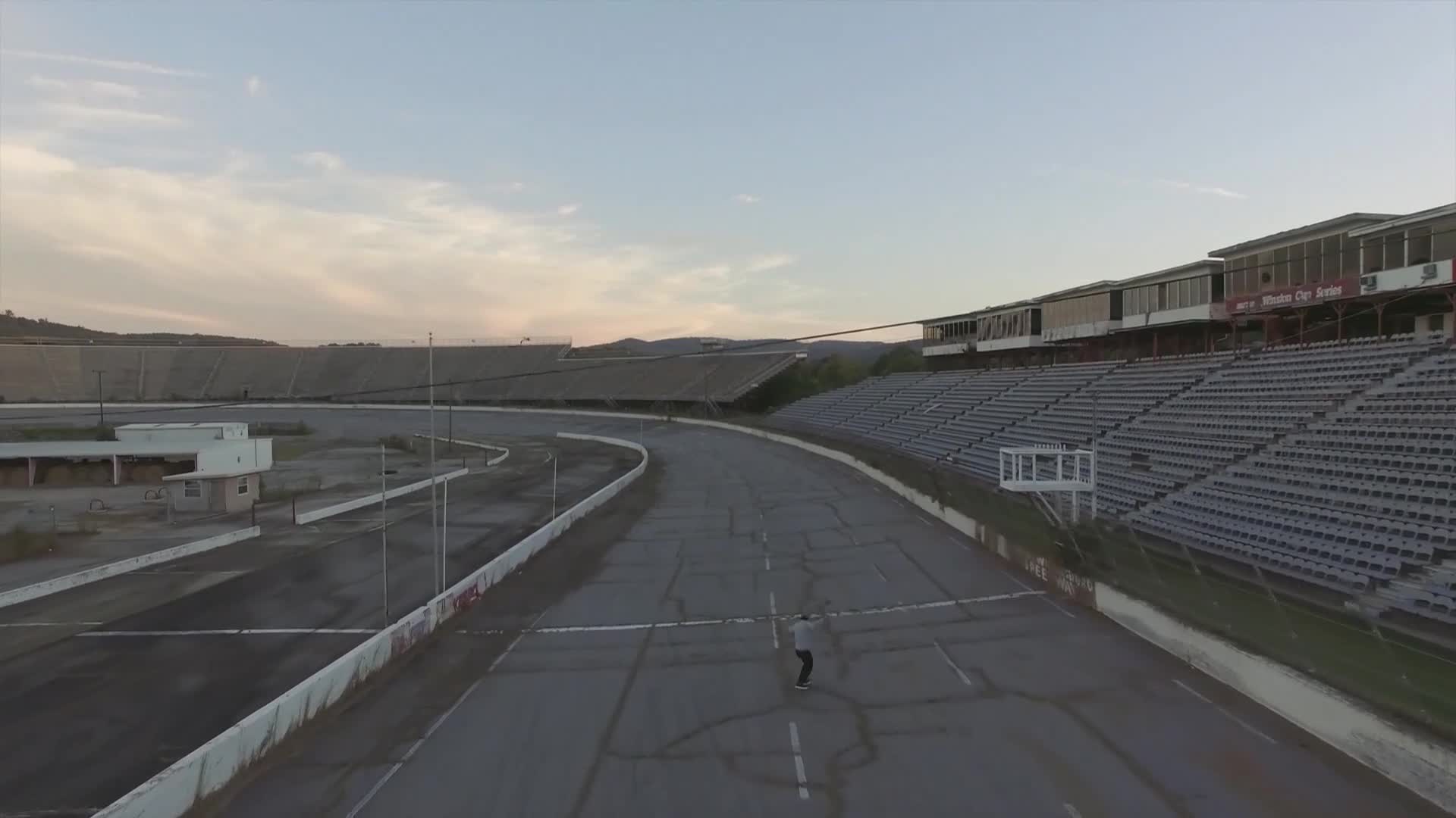 A lone figure stands on the cracked asphalt of a deserted racetrack. The grandstands are empty, and the sky is overcast.
