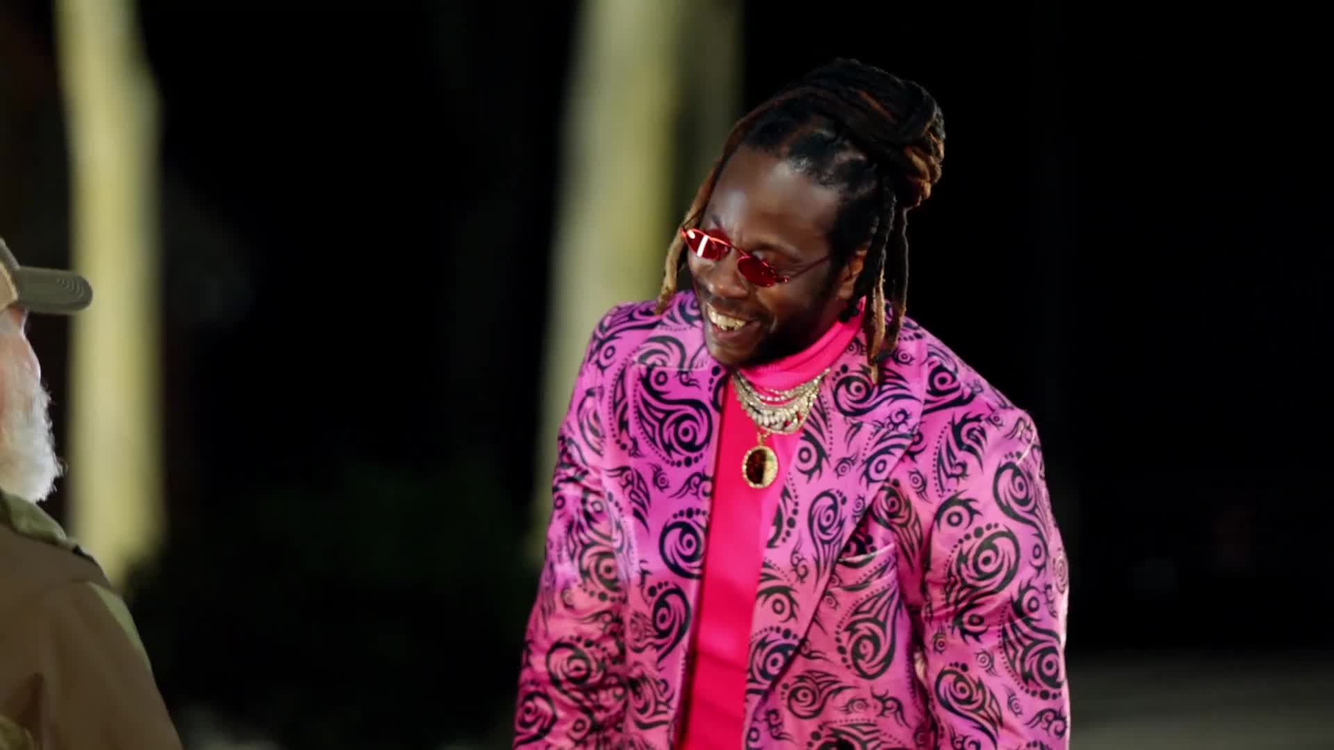 A man in a vibrant pink paisley suit and matching turtleneck laughs, his dreadlocks pulled back. He's talking to someone off-camera, their face mostly obscured.