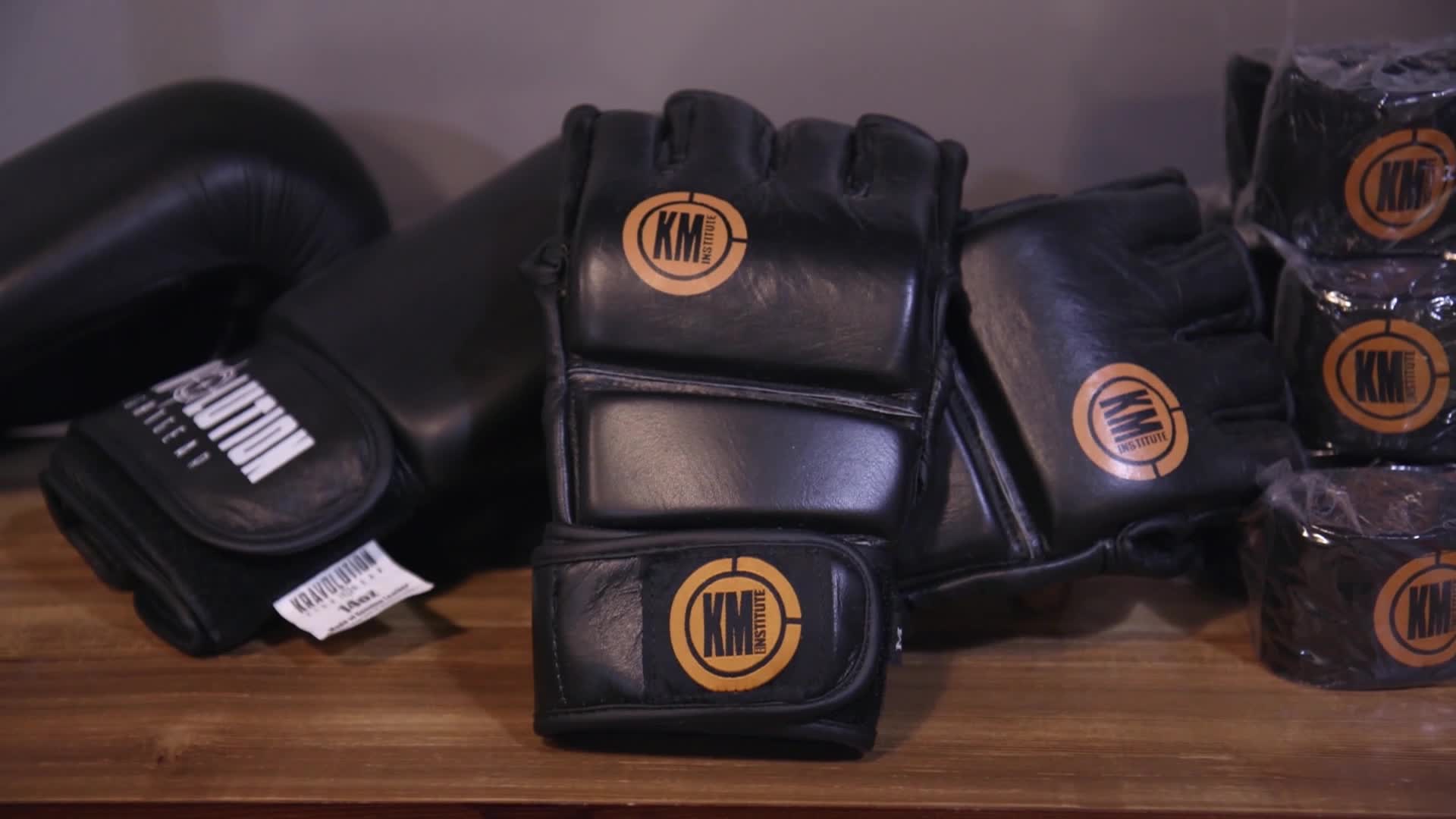 Black MMA gloves with a gold "KM Institute" logo sit on a wooden surface. To the left, another pair of black gloves, labeled "Kravolution," rests beside them.