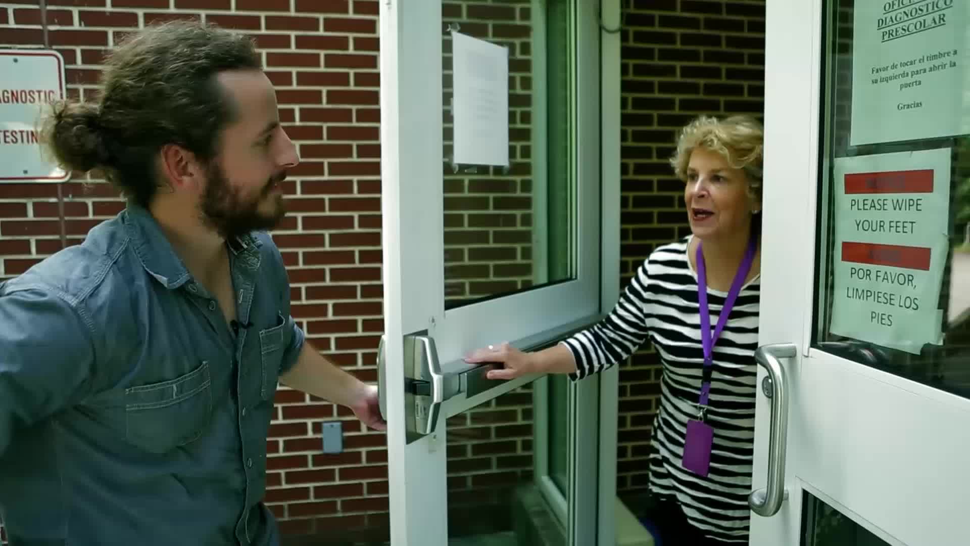 A woman in a striped shirt opens a glass door, her hand on the push bar. A man with his hair in a bun turns to face her, his own hand reaching for the door.