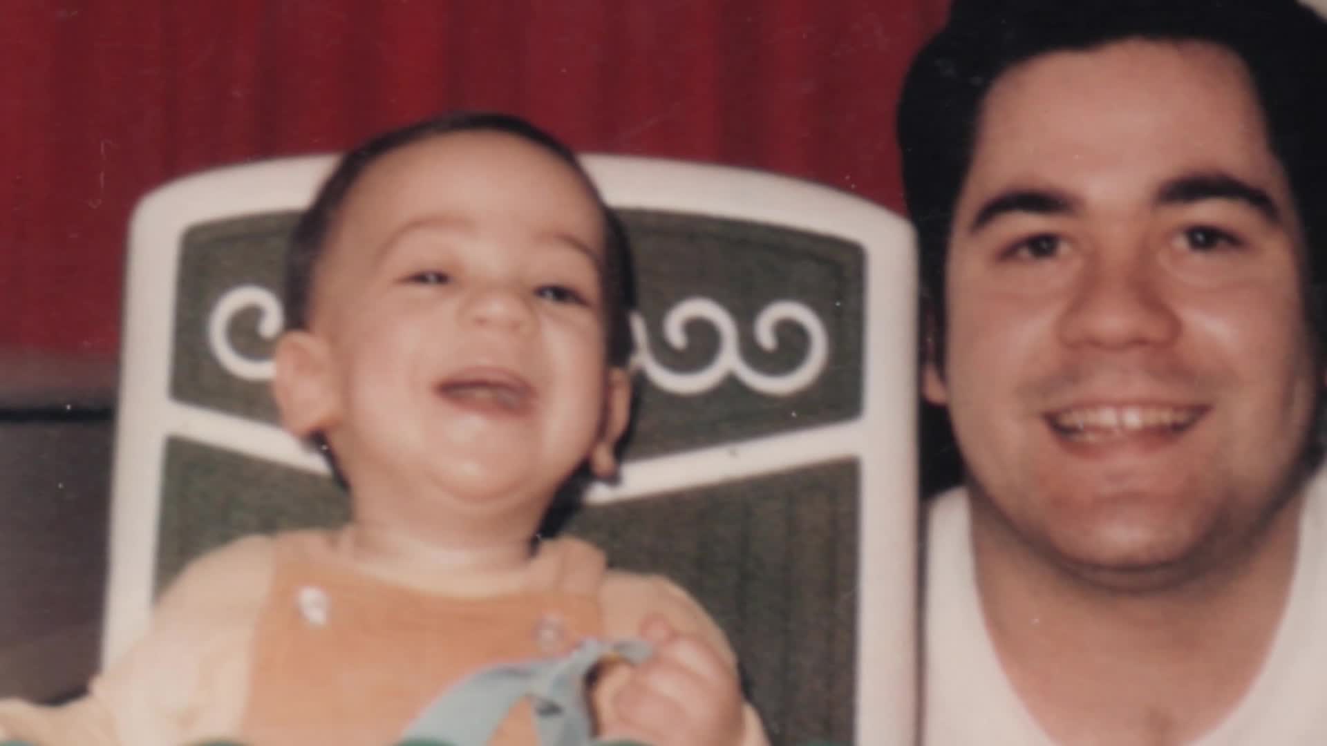 A baby with a wide grin is propped up in a high chair. Beside him, a man smiles warmly.