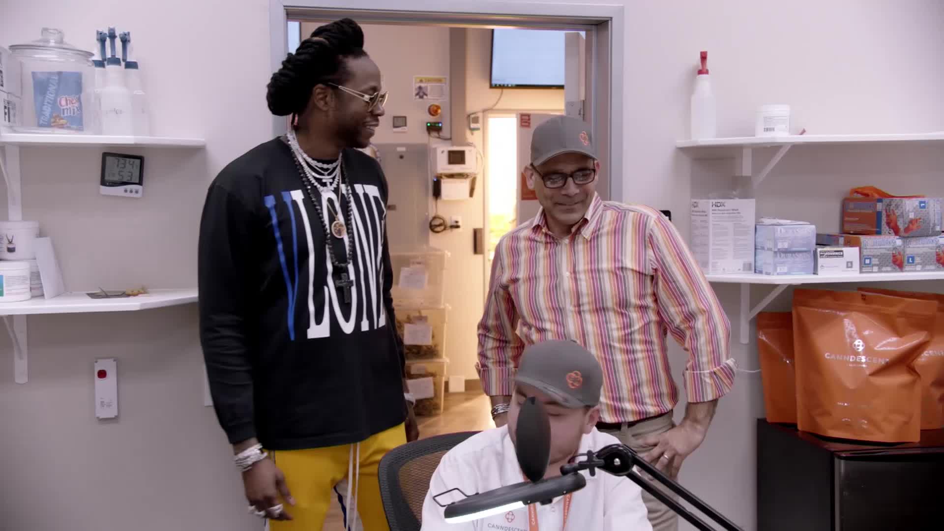 A man in a black shirt and yellow pants stands talking with another man in a striped shirt. A third person is seated at a microphone in front of them. This is happening inside a room that looks like a lab or storage area.