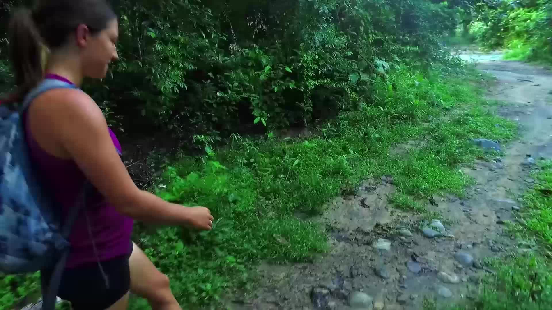 A woman in a purple tank top and black shorts walks along a muddy path, her backpack slung over her shoulder. Lush green foliage crowds the edges of the trail, hinting at a humid, tropical environment.