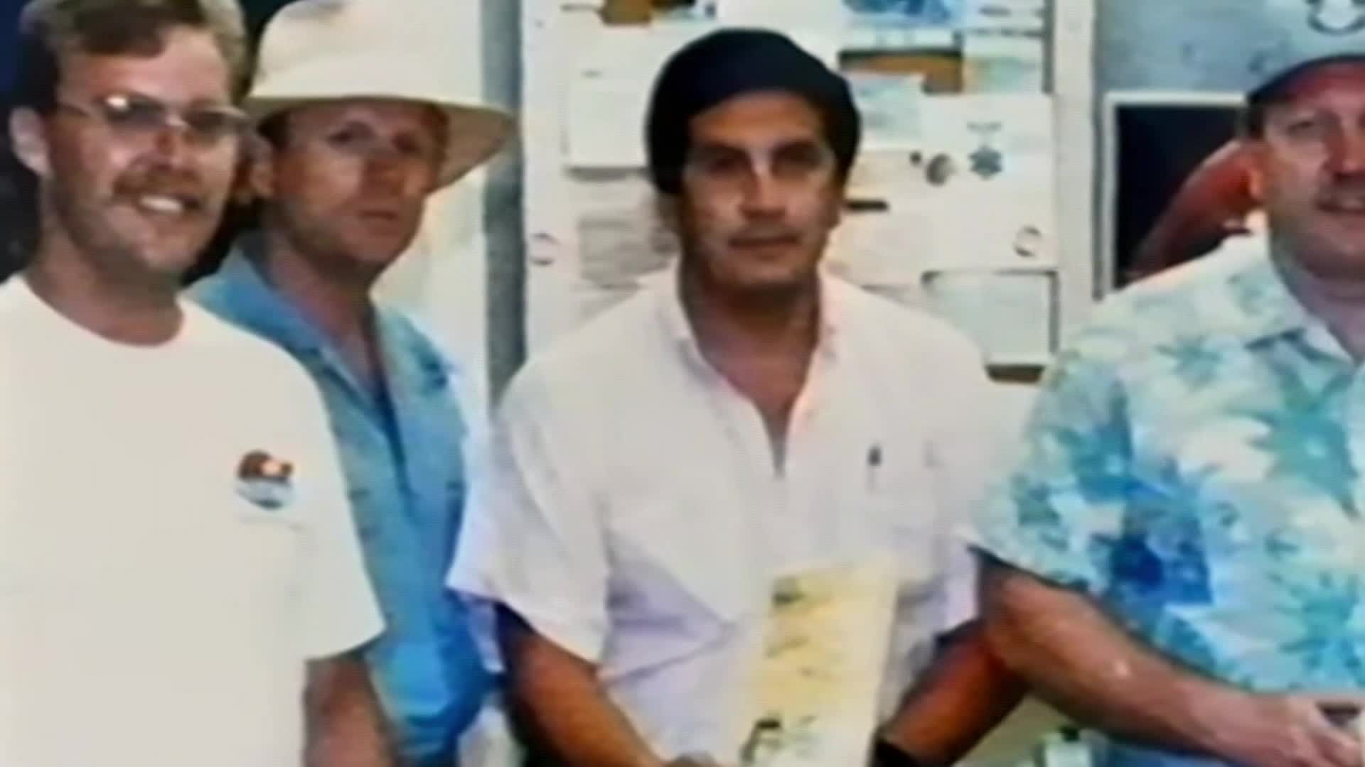 Four men stand together, their faces turned towards the camera. One man in a white t-shirt with glasses smiles, while another in a light blue shirt and a wide-brimmed hat looks off to the side. A third man in a white polo shirt holds a document, and the fourth, wearing a colorful floral shirt, stands to his right.