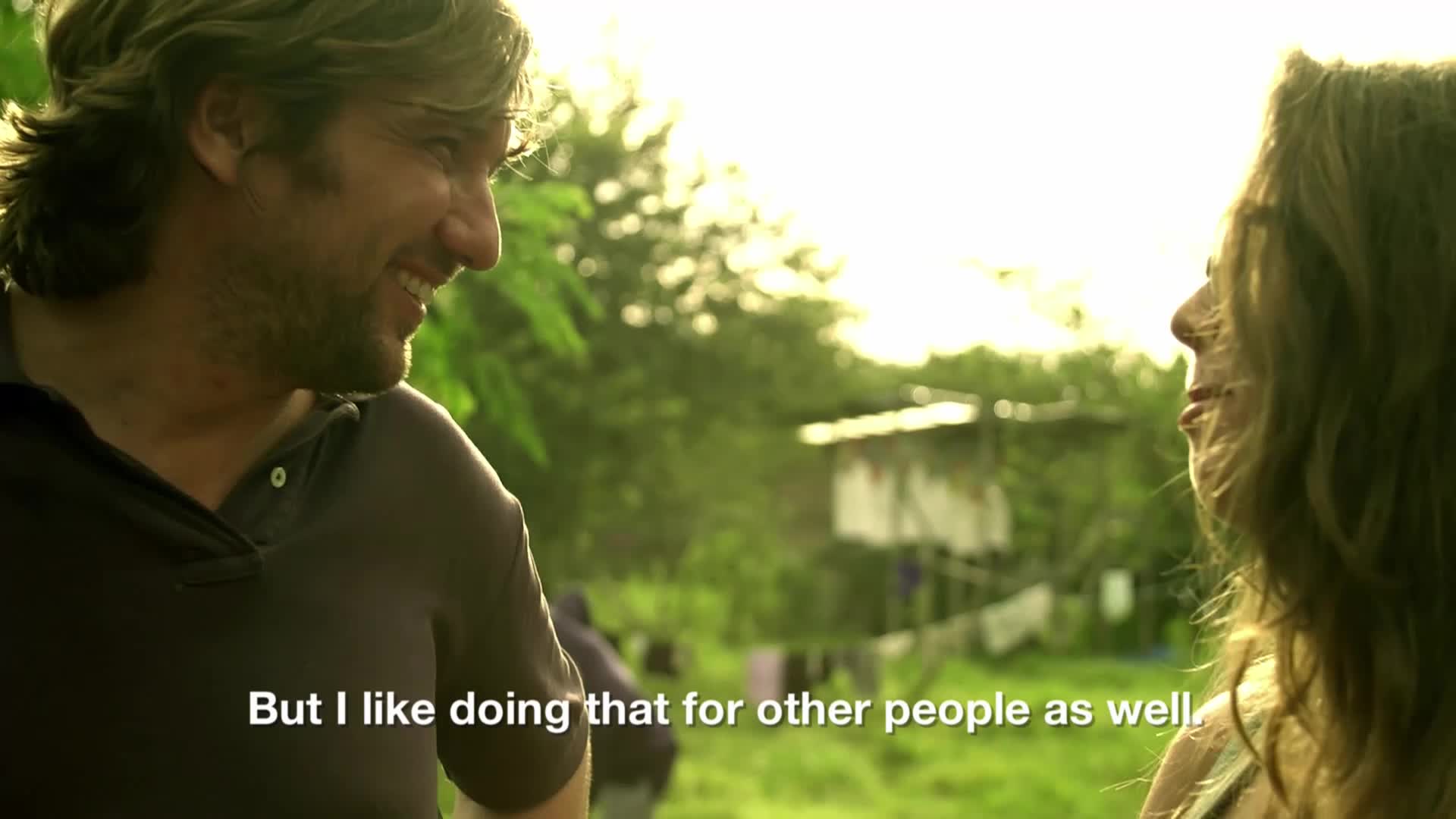 A man with shaggy hair smiles, his gaze directed off-camera. Behind him, a woman's profile is visible, her hair catching the light. The background is a blur of green foliage and a simple structure with clothes hanging to dry.
