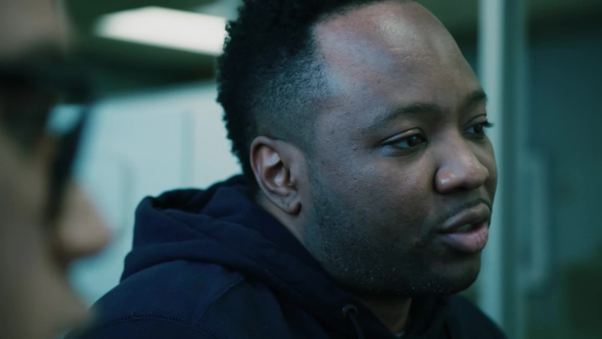 A man with a sharp haircut speaks, his dark hoodie pulled up. He's talking to someone just out of frame, the conversation likely for a VICE TV segment.