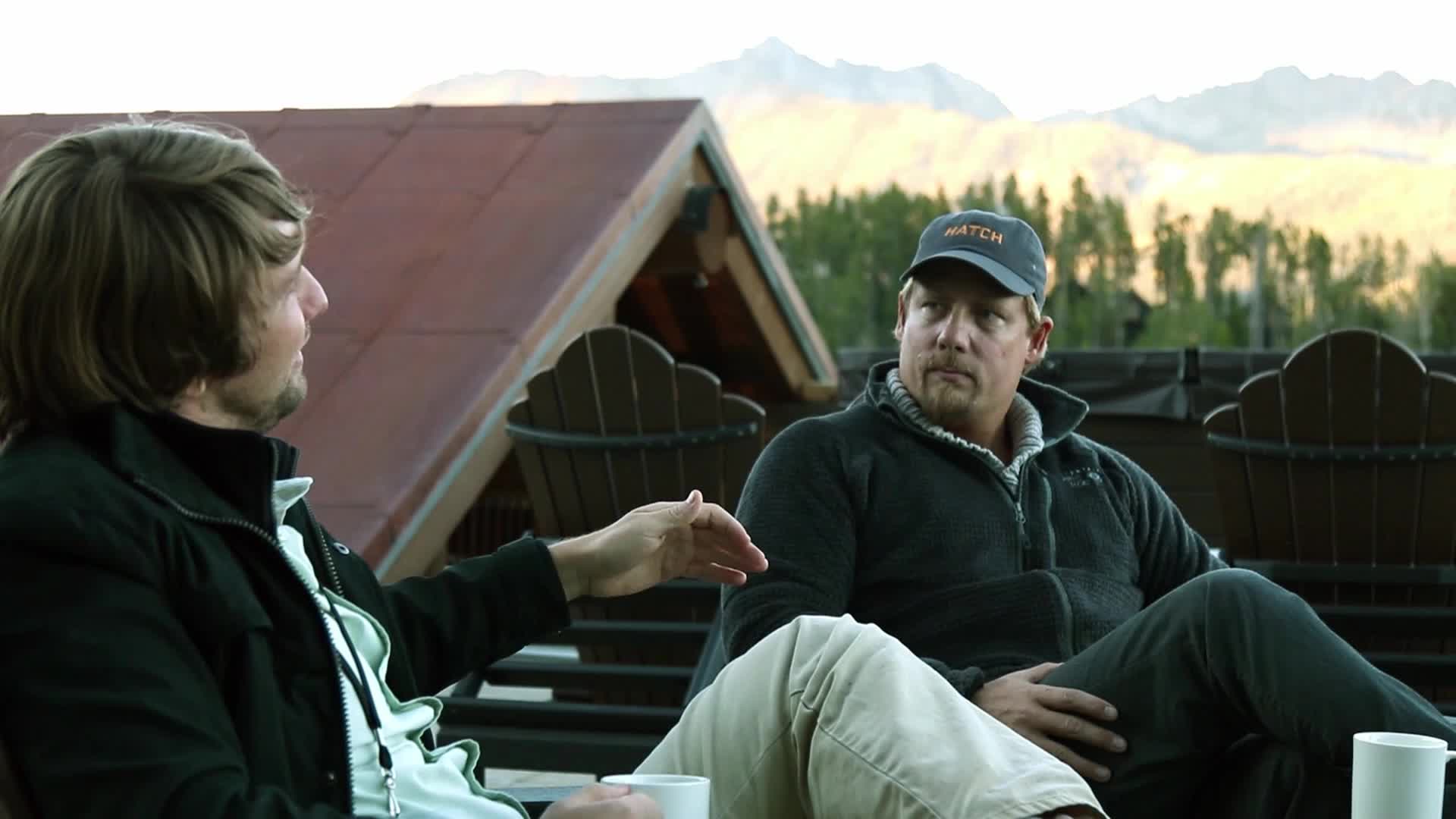 Two men sit outdoors, a conversation unfolding between them. One gestures with his hand, his face turned towards the other.