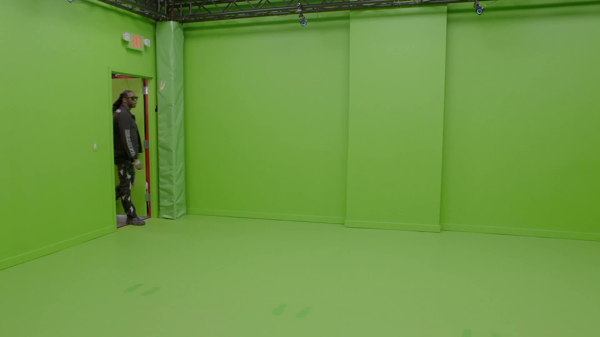 A man stands in a doorway, framed by a bright green wall. He's wearing a dark jacket and patterned pants, looking out into the empty studio space.