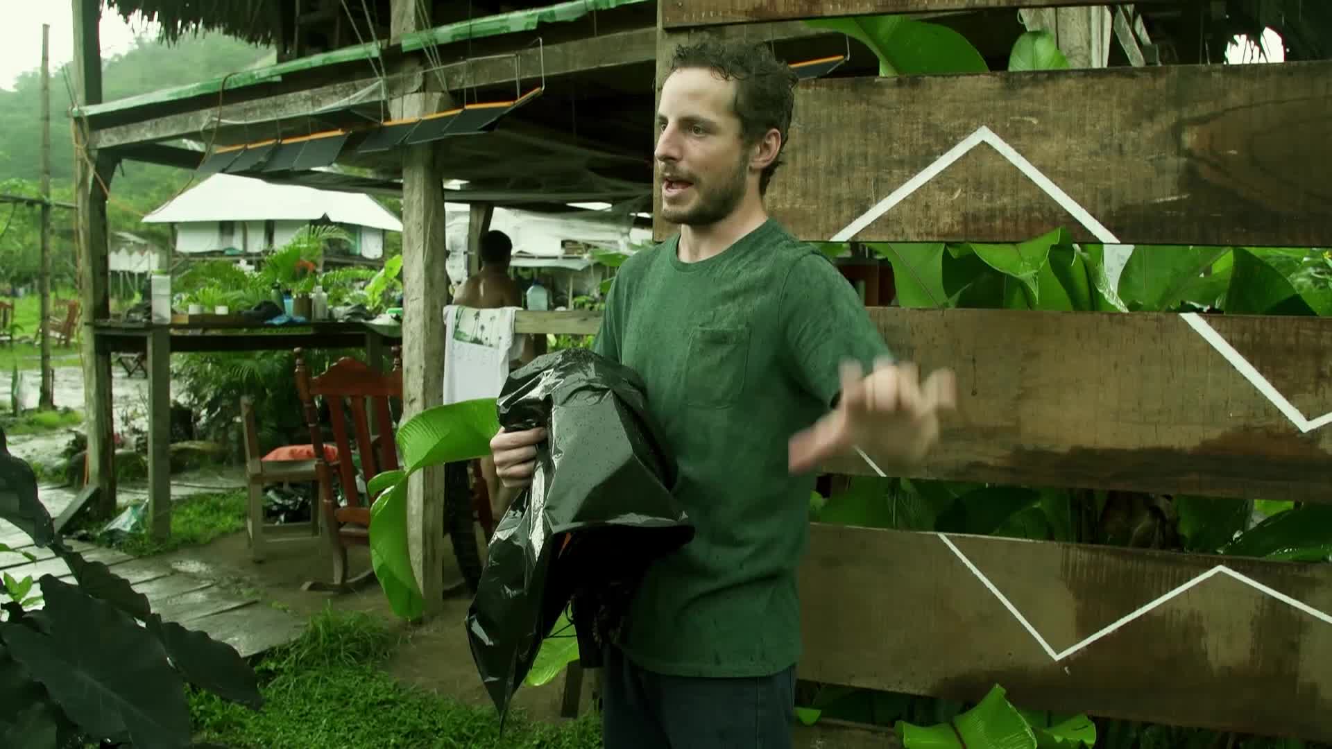 A man in a green t-shirt gestures with his hands while holding a crumpled black bag. He's speaking in front of a wooden fence adorned with white geometric patterns.