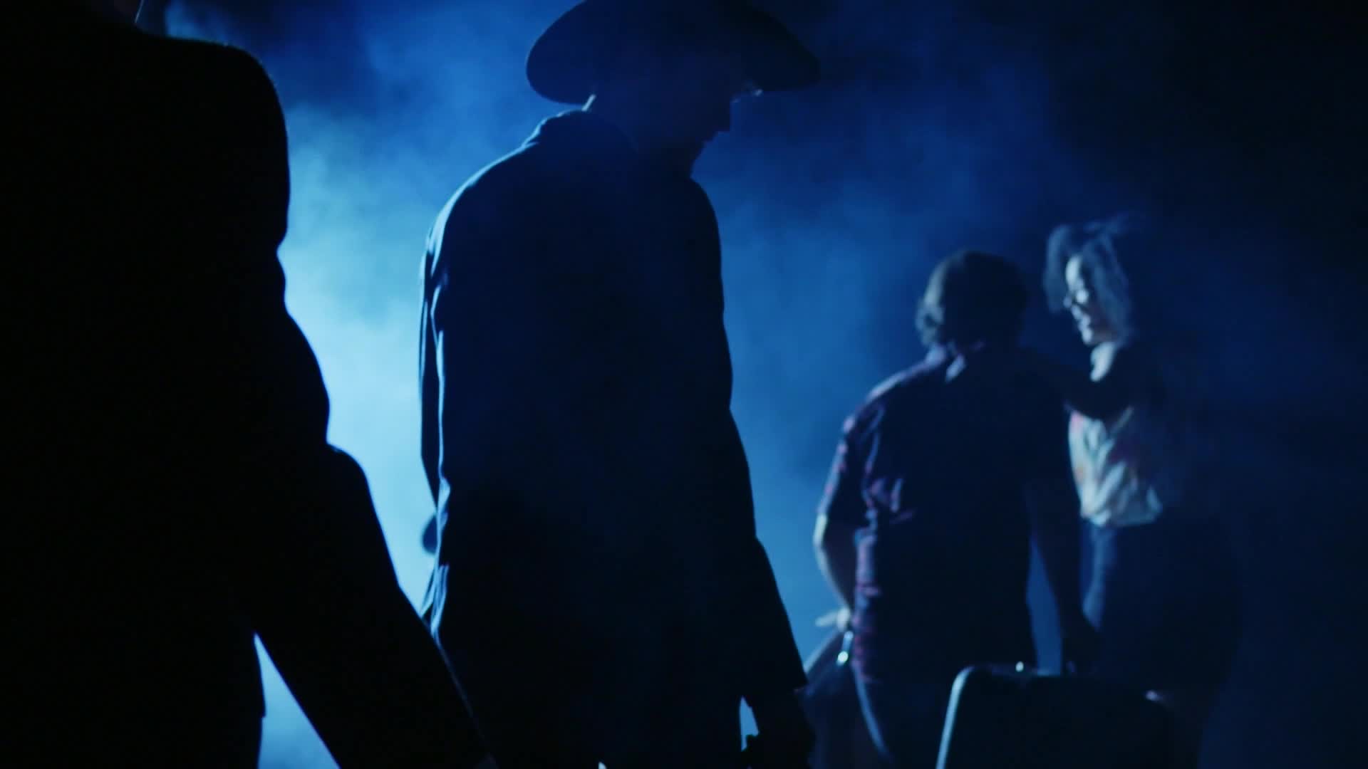 A man in a cowboy hat stands in silhouette, bathed in blue light. Behind him, two figures huddle close, their faces obscured by shadow and haze.