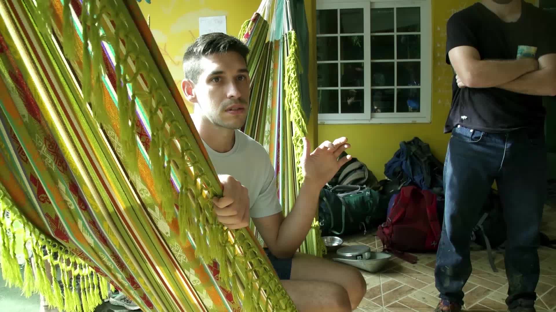 A man in a light gray t-shirt gestures with his right hand while holding onto a brightly colored hammock. Behind him, another man in a black t-shirt and jeans stands with his arms crossed, surrounded by backpacks.