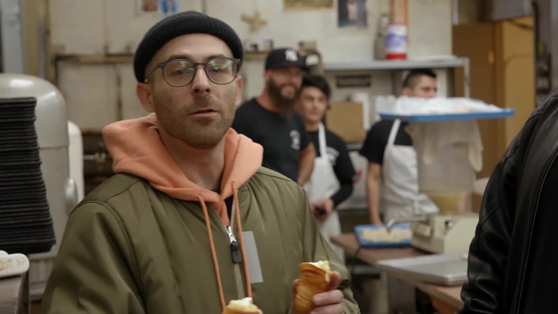 A man in a beanie and glasses holds two pastries, looking directly ahead. Behind him, other men in aprons work in a bakery.