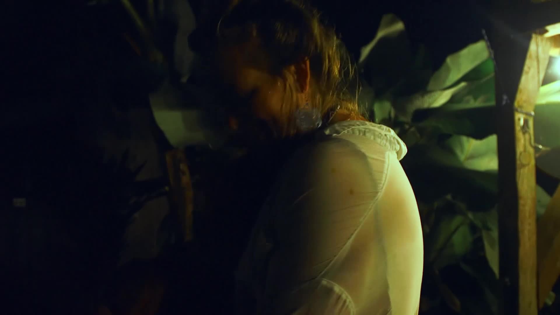 A woman in a sheer white dress leans forward, her face obscured by shadow. She's in a dimly lit space, surrounded by what looks like large, dark leaves.