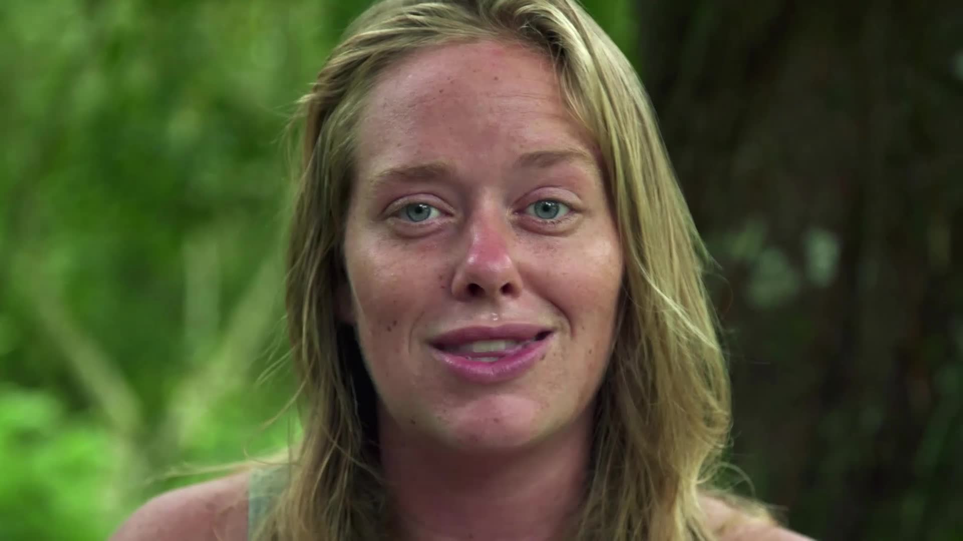 A young woman with blonde hair and blue eyes looks directly at me, her face flushed from the sun. She's speaking, her lips parted slightly, with a backdrop of lush green foliage.
