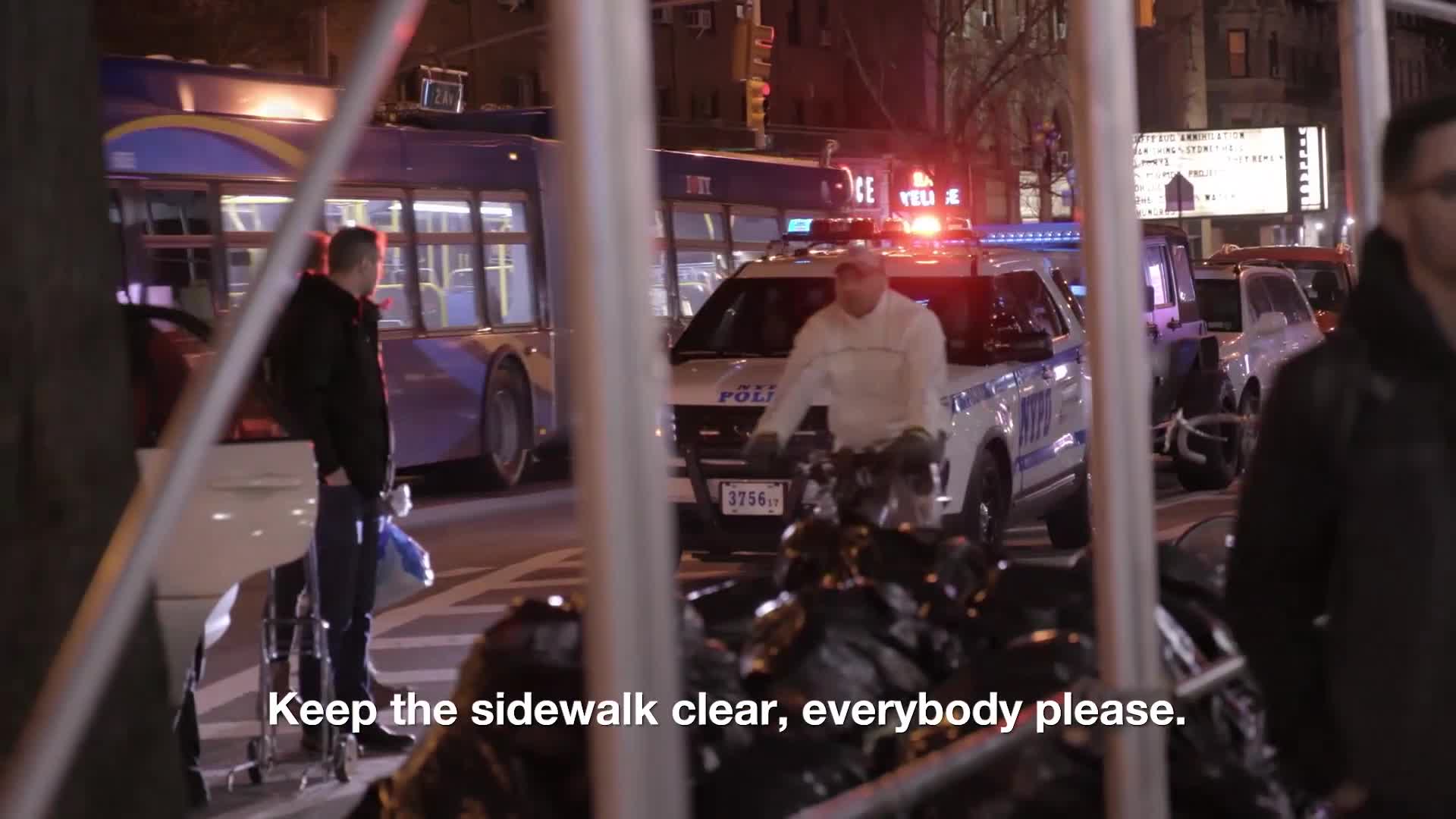 A police car with flashing lights is parked on the street, and a man in a white jacket is standing next to it. A bus is stopped behind the police car, and people are walking on the sidewalk.