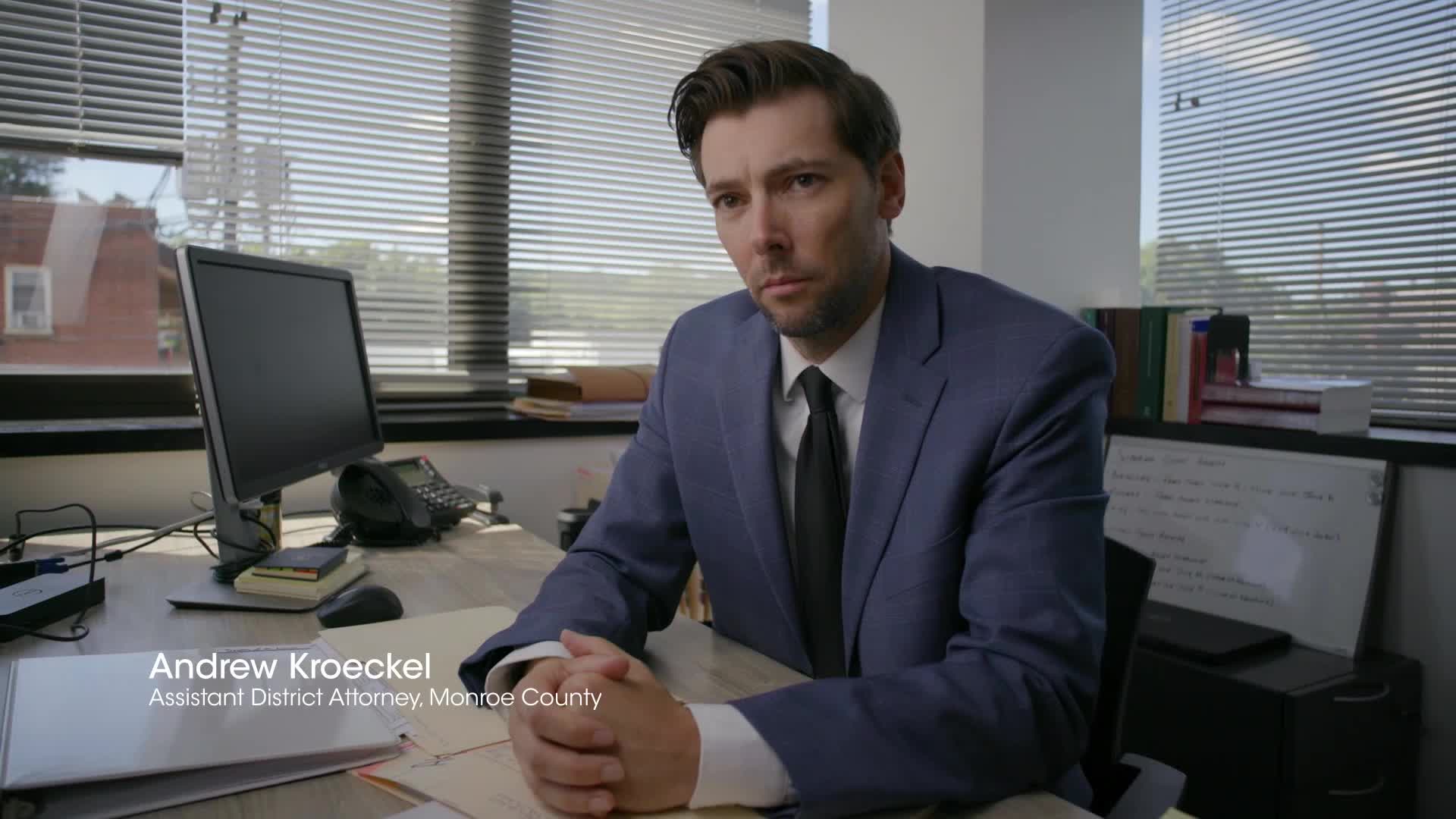 Andrew Kroeckel, an Assistant District Attorney from Monroe County, looks directly ahead. His hands are clasped on the desk in front of him.
