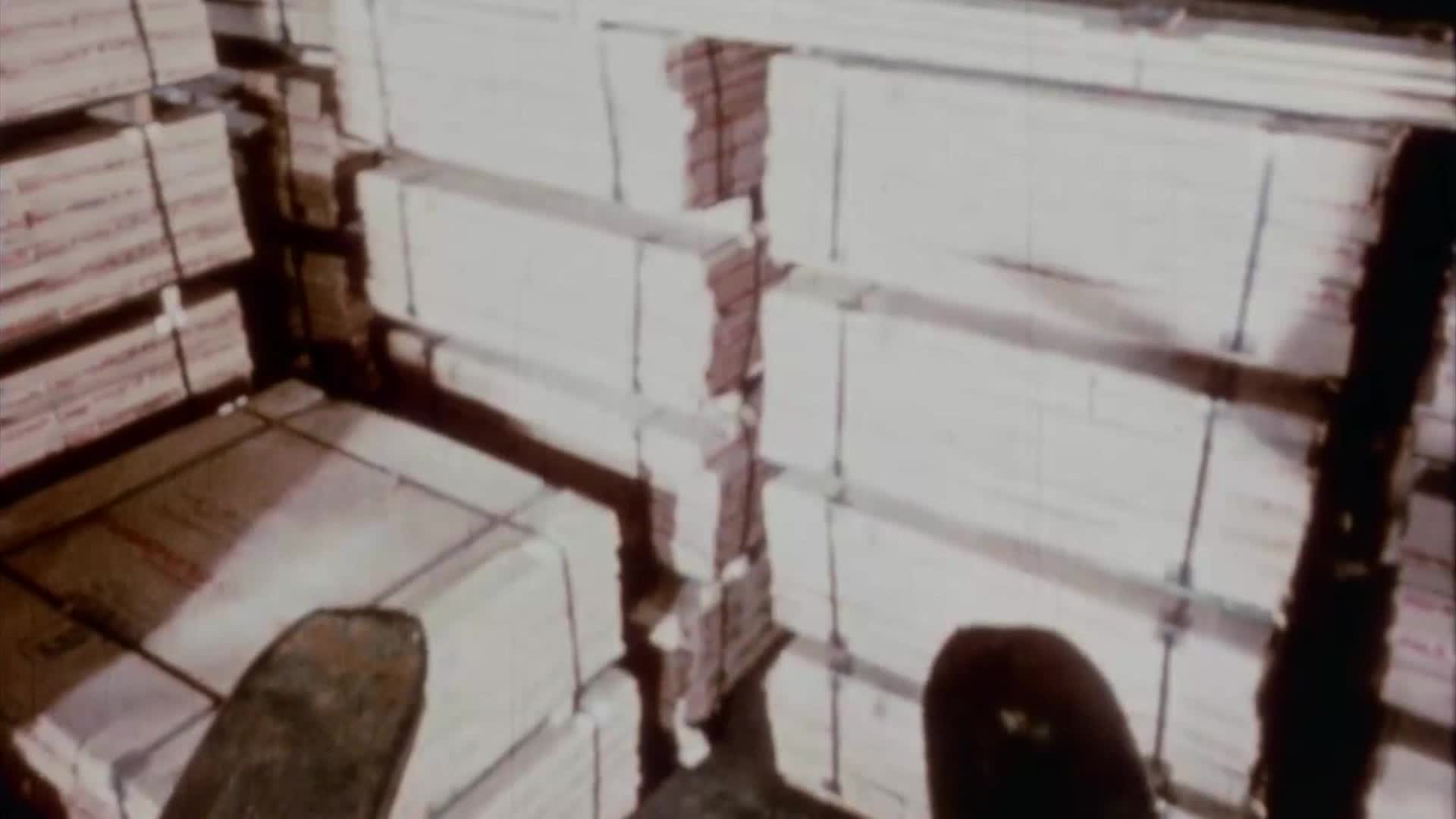 Stacks of white paper bundles fill the warehouse. A dark, curved object, possibly a shoe, rests on the floor in the foreground.