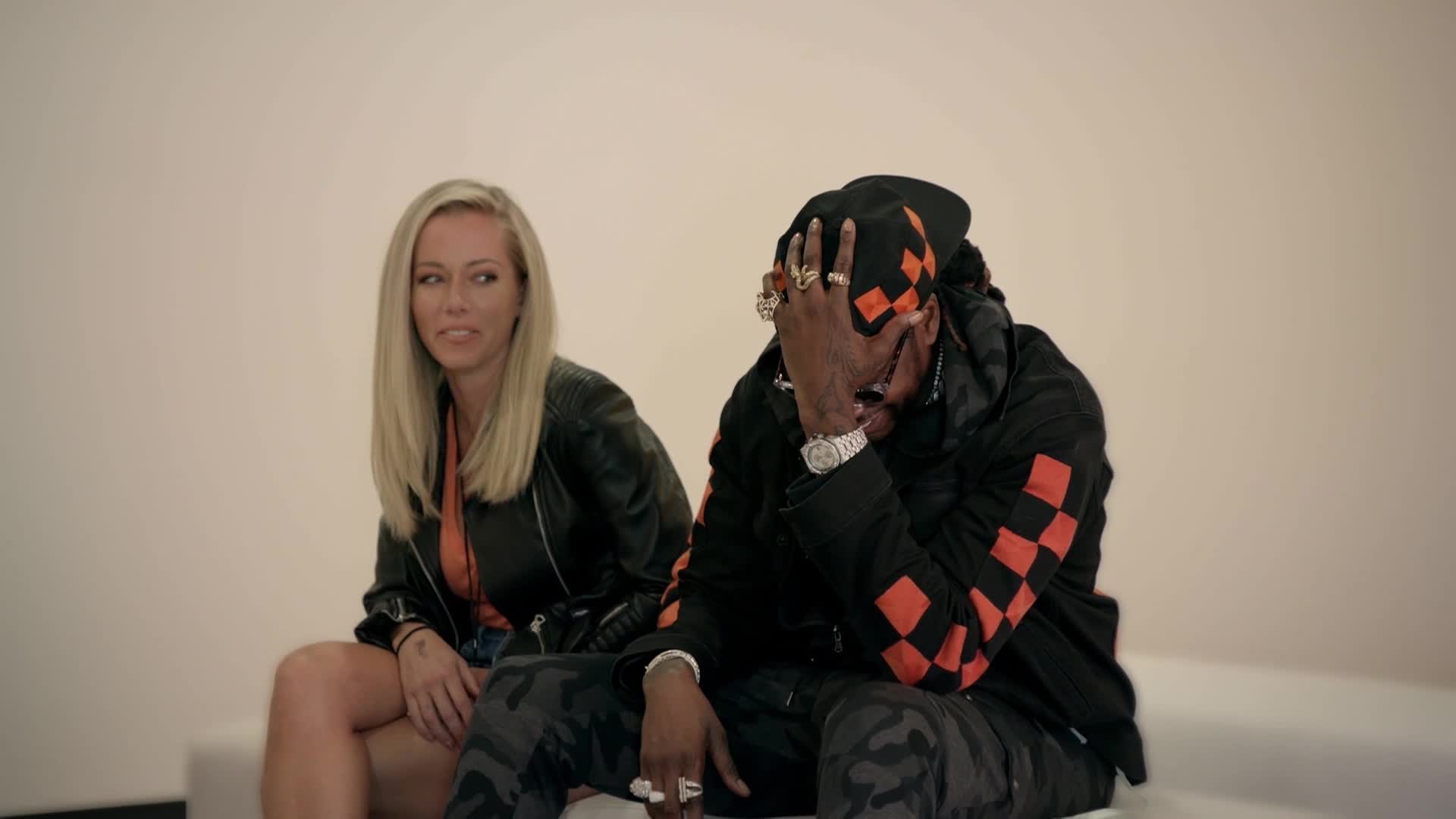 A woman with blonde hair sits to the left, looking off-camera. Beside her, a man in a black hoodie with orange checkered accents covers his face with his hand.