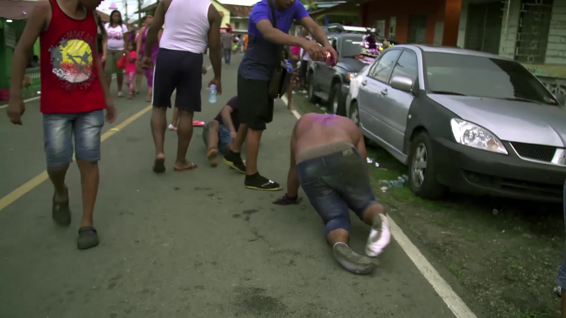 A man is doing push-ups on the street, his back dusted with purple powder. People are walking past, some watching the man as he moves.