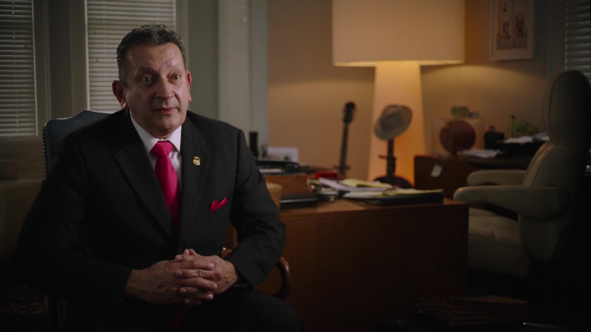 A man in a black suit and bright pink tie speaks from his office. Papers and a basketball sit on his desk behind him.