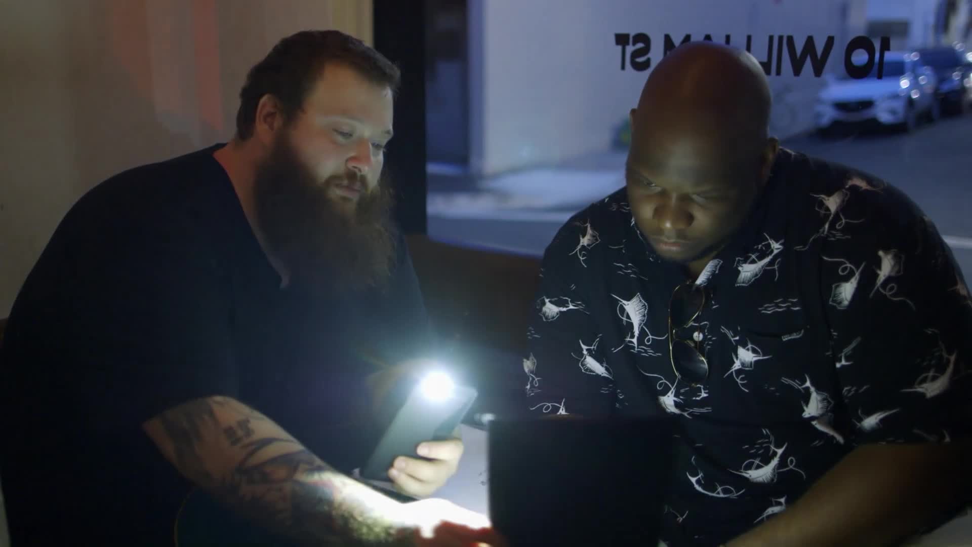 Two men are hunched over a table, illuminated by a bright phone light and a laptop screen. Outside the window, cars pass by on a city street at night.