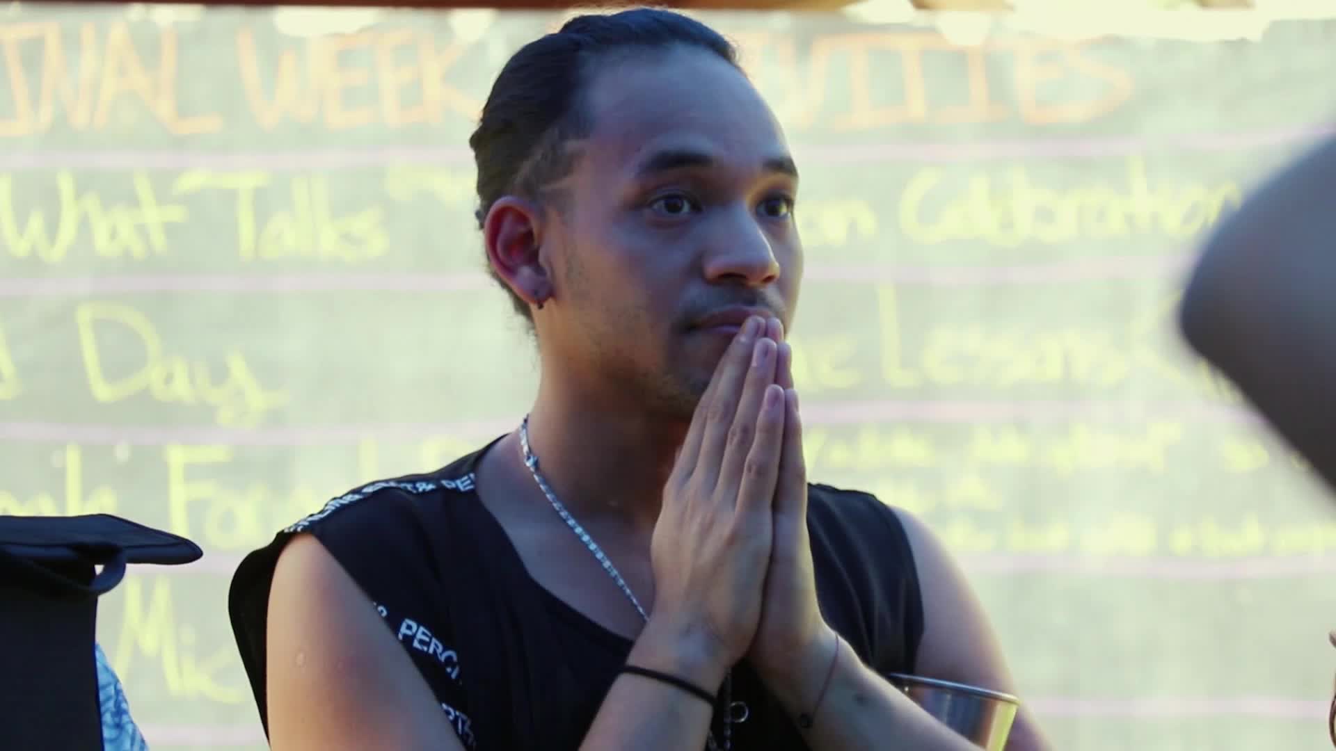 He clasps his hands together, eyes wide, as if listening intently to something beyond the frame. The words "Festival Week" and "What Talks" are scrawled on a board behind him.